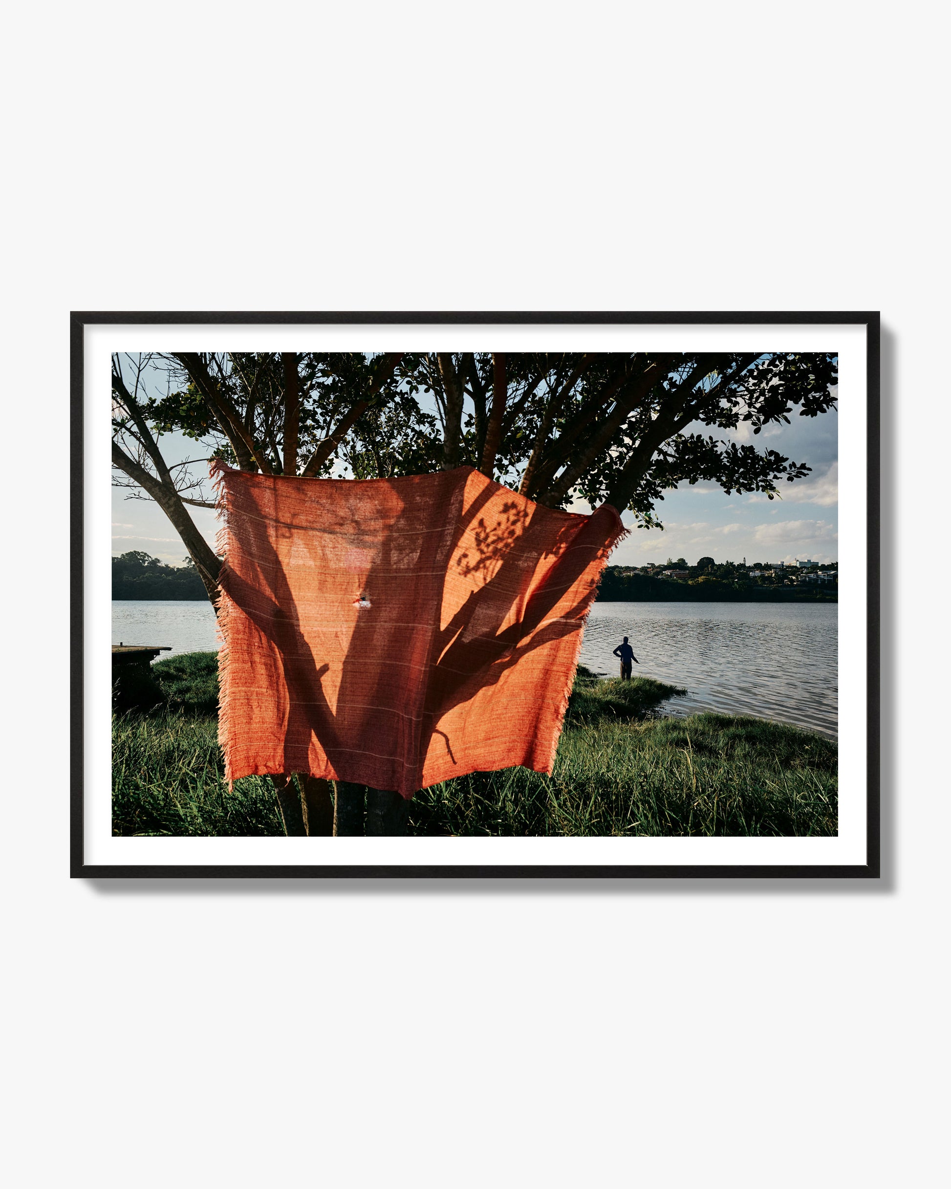 Fine art street photograph of a vibrant orange cloth hanging from a tree, with a tranquil lake and a fisherman in the background. Archival pigment print, black frame.