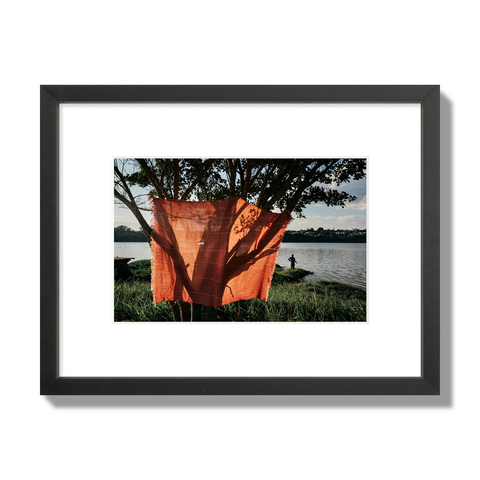 Fine art street photograph of a vibrant orange cloth hanging from a tree, with a tranquil lake and a fisherman in the background. Archival pigment print, small size, black frame.