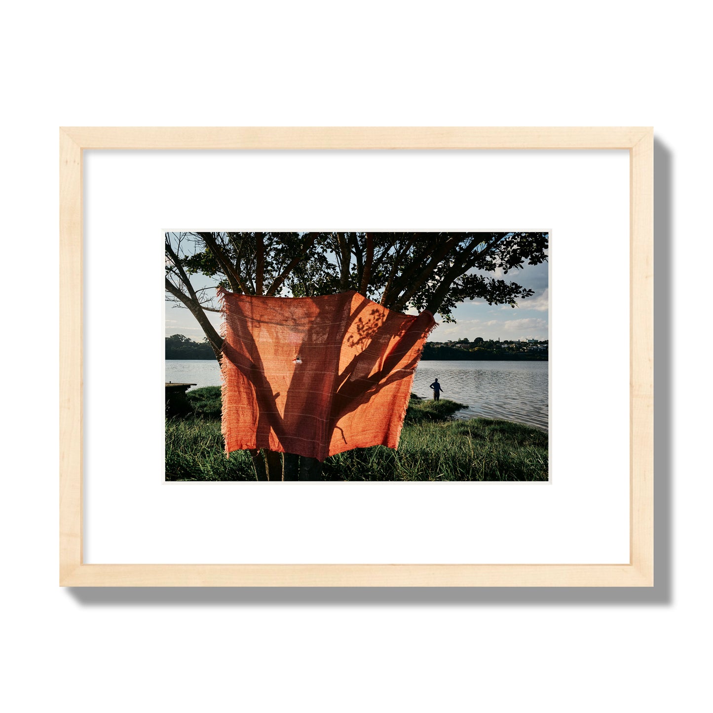 Fine art street photograph of a vibrant orange cloth hanging from a tree, with a tranquil lake and a fisherman in the background. Archival pigment print, small size, natural wood frame.
