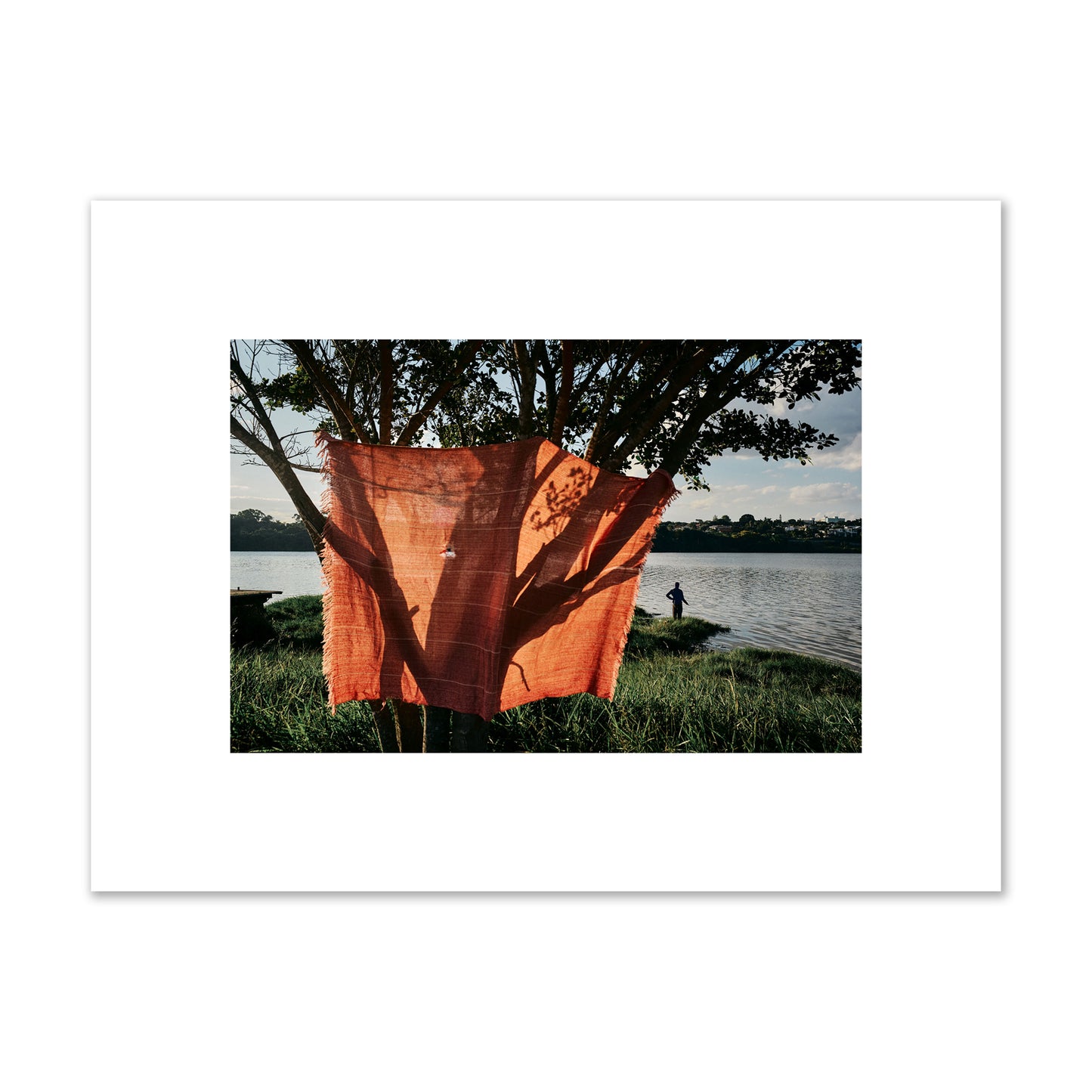 Fine art street photograph of a vibrant orange cloth hanging from a tree, with a tranquil lake and a fisherman in the background. Archival pigment print, small size, unframed.