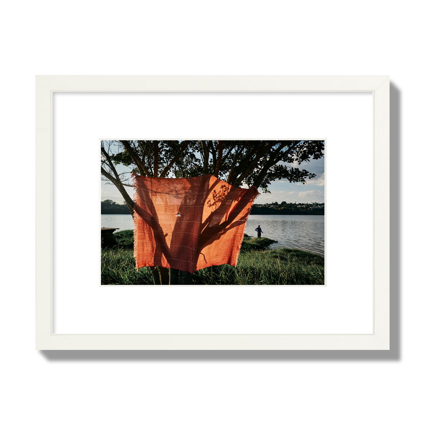 Fine art street photograph of a vibrant orange cloth hanging from a tree, with a tranquil lake and a fisherman in the background. Archival pigment print, small size, white frame.