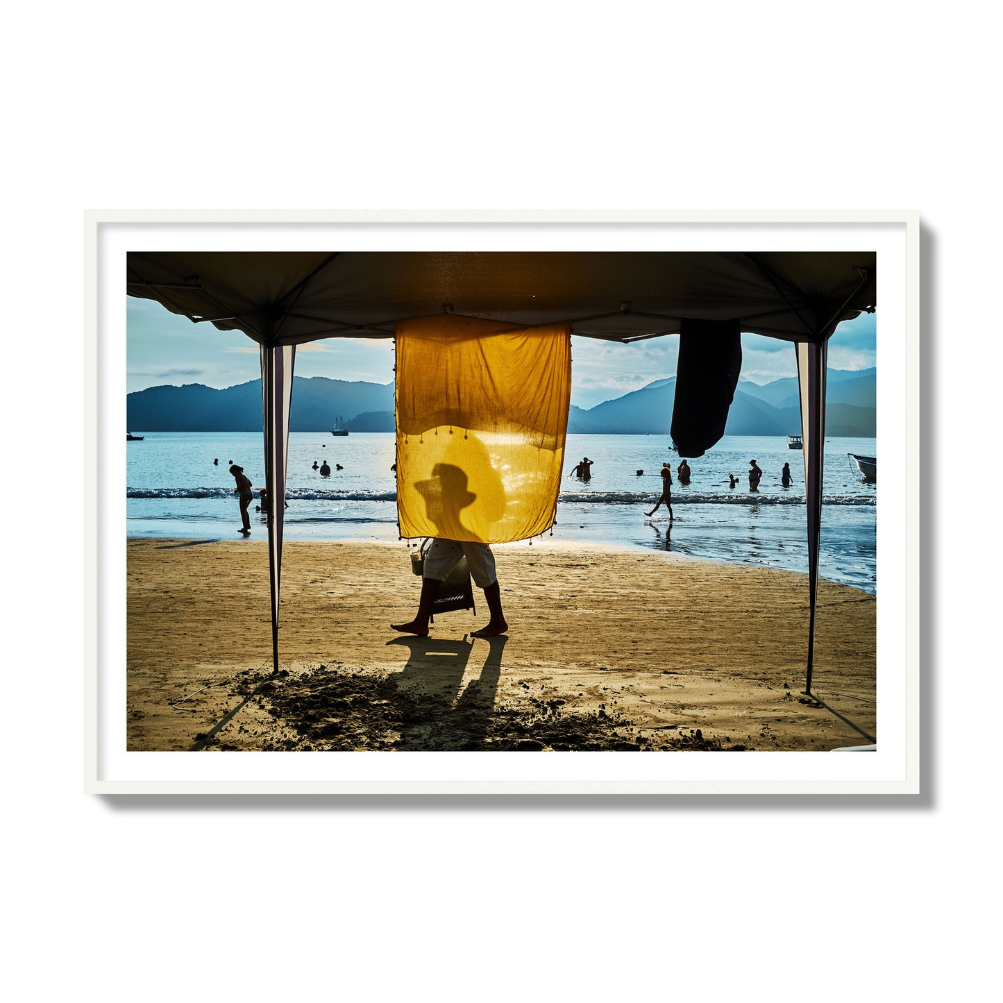 Fine art street photograph of a crowded beach scene with a vibrant yellow flap hanging from a tent, sunlight filtering through and silhouetting a man’s face. Archival pigment print, collector size, white frame.