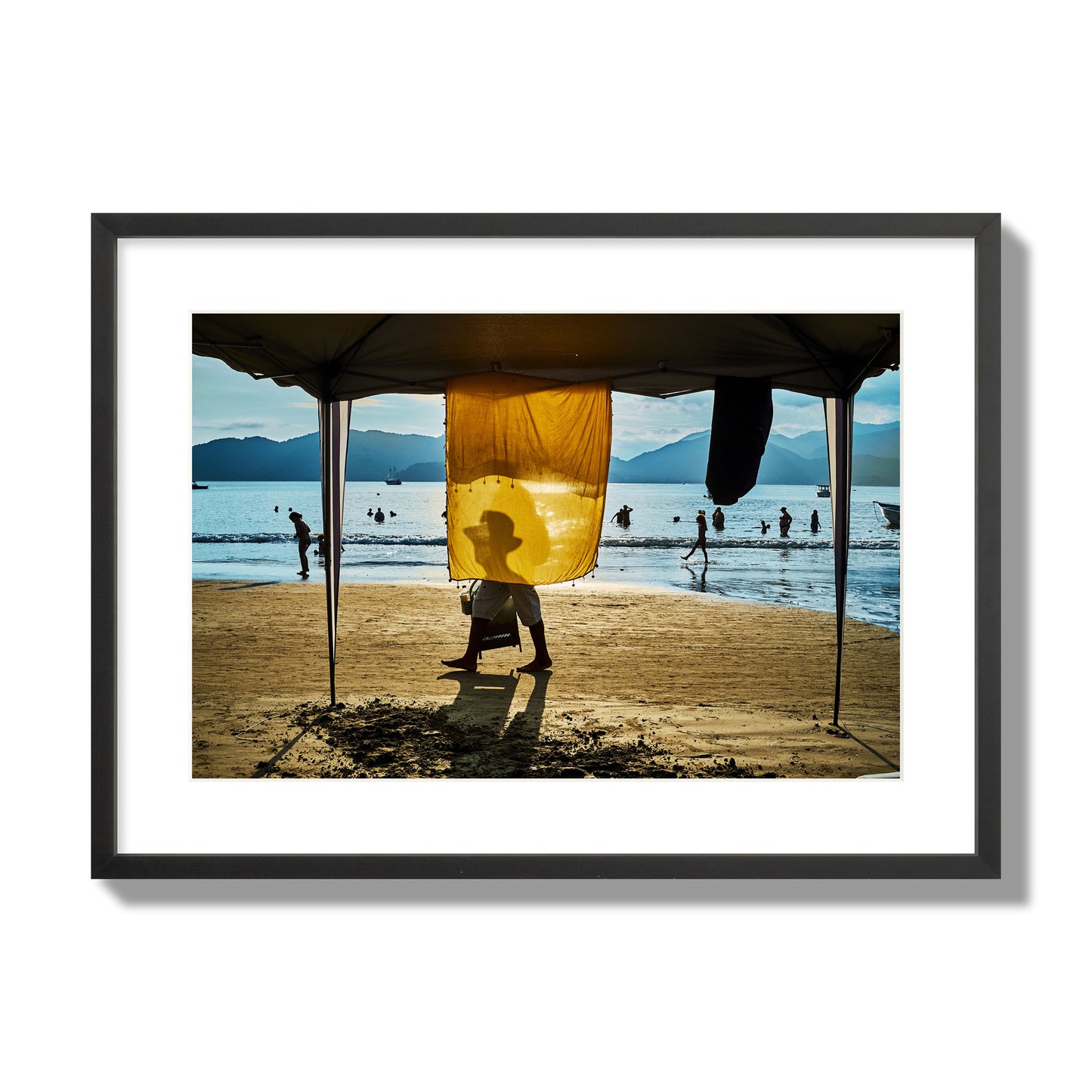 Fine art street photograph of a crowded beach scene with a vibrant yellow flap hanging from a tent, sunlight filtering through and silhouetting a man’s face. Archival pigment print, large size, black frame.