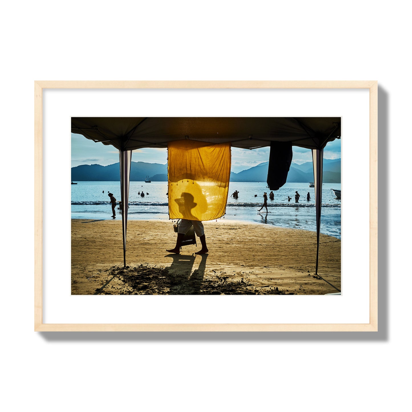 Fine art street photograph of a crowded beach scene with a vibrant yellow flap hanging from a tent, sunlight filtering through and silhouetting a man’s face. Archival pigment print, large size, natural wood frame.