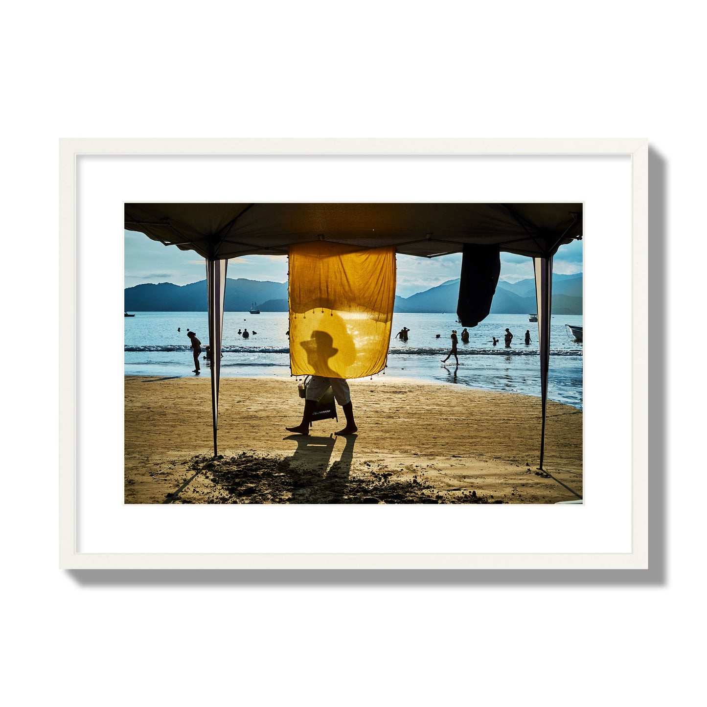 Fine art street photograph of a crowded beach scene with a vibrant yellow flap hanging from a tent, sunlight filtering through and silhouetting a man’s face. Archival pigment print, large size, white frame.
