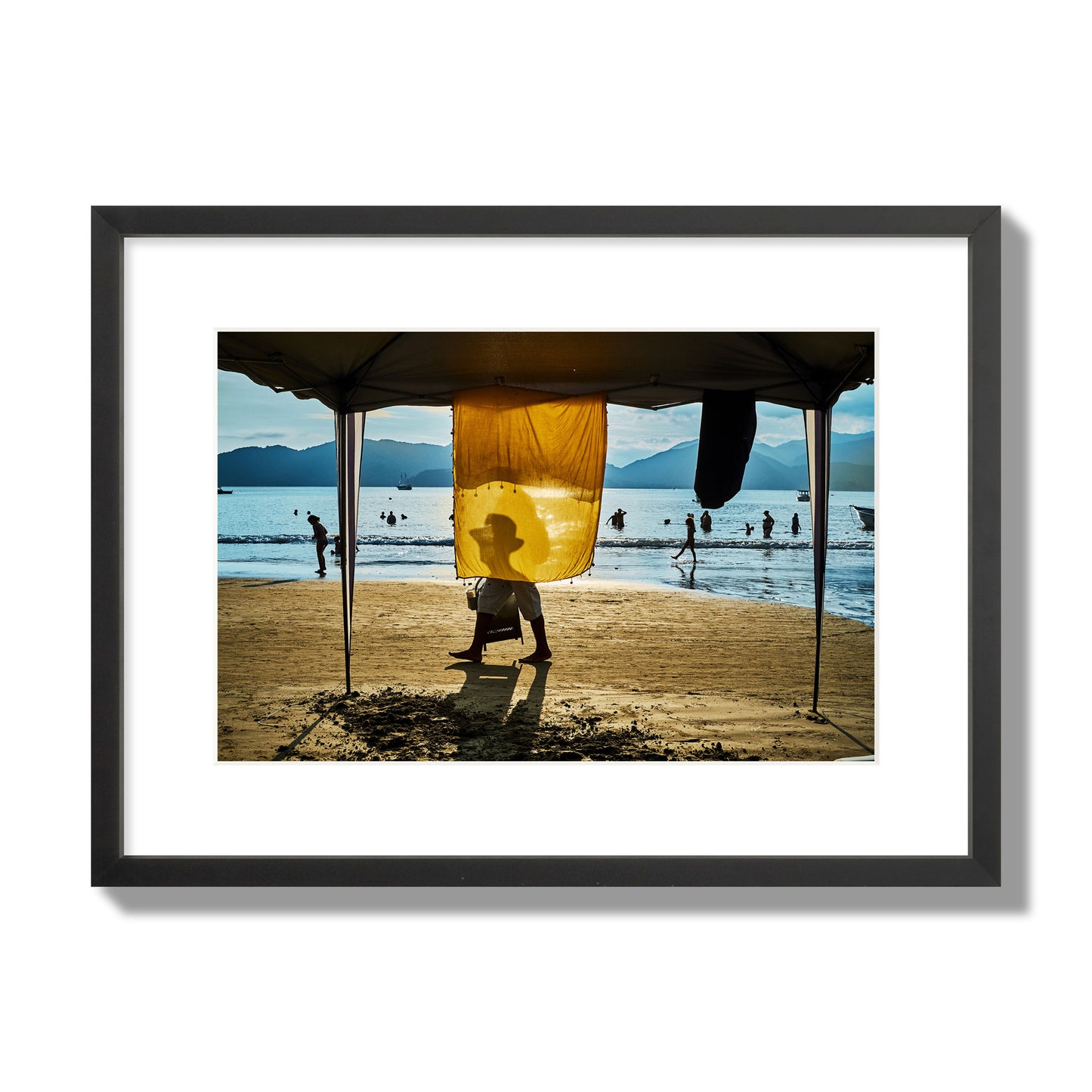 Fine art street photograph of a crowded beach scene with a vibrant yellow flap hanging from a tent, sunlight filtering through and silhouetting a man’s face. Archival pigment print, medium size, black frame.