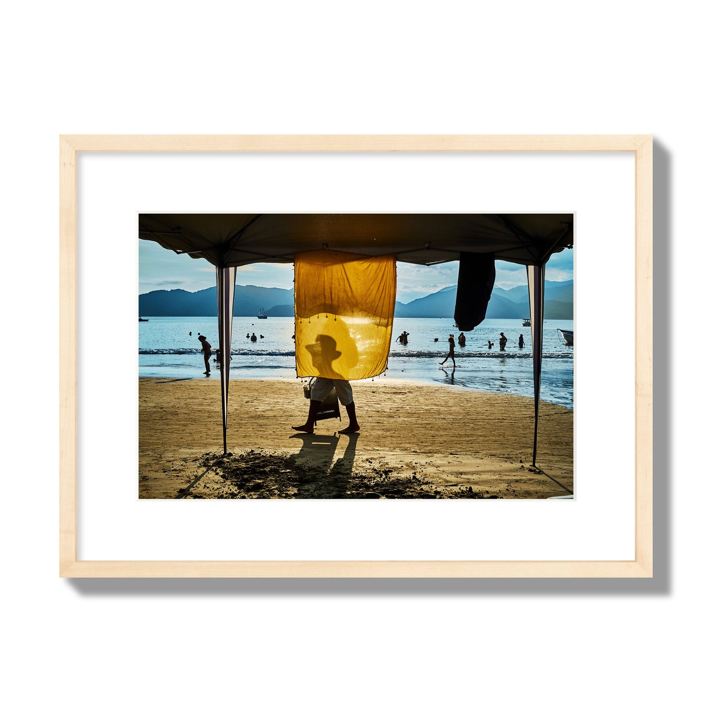 Fine art street photograph of a crowded beach scene with a vibrant yellow flap hanging from a tent, sunlight filtering through and silhouetting a man’s face. Archival pigment print, medium size, natural wood frame.
