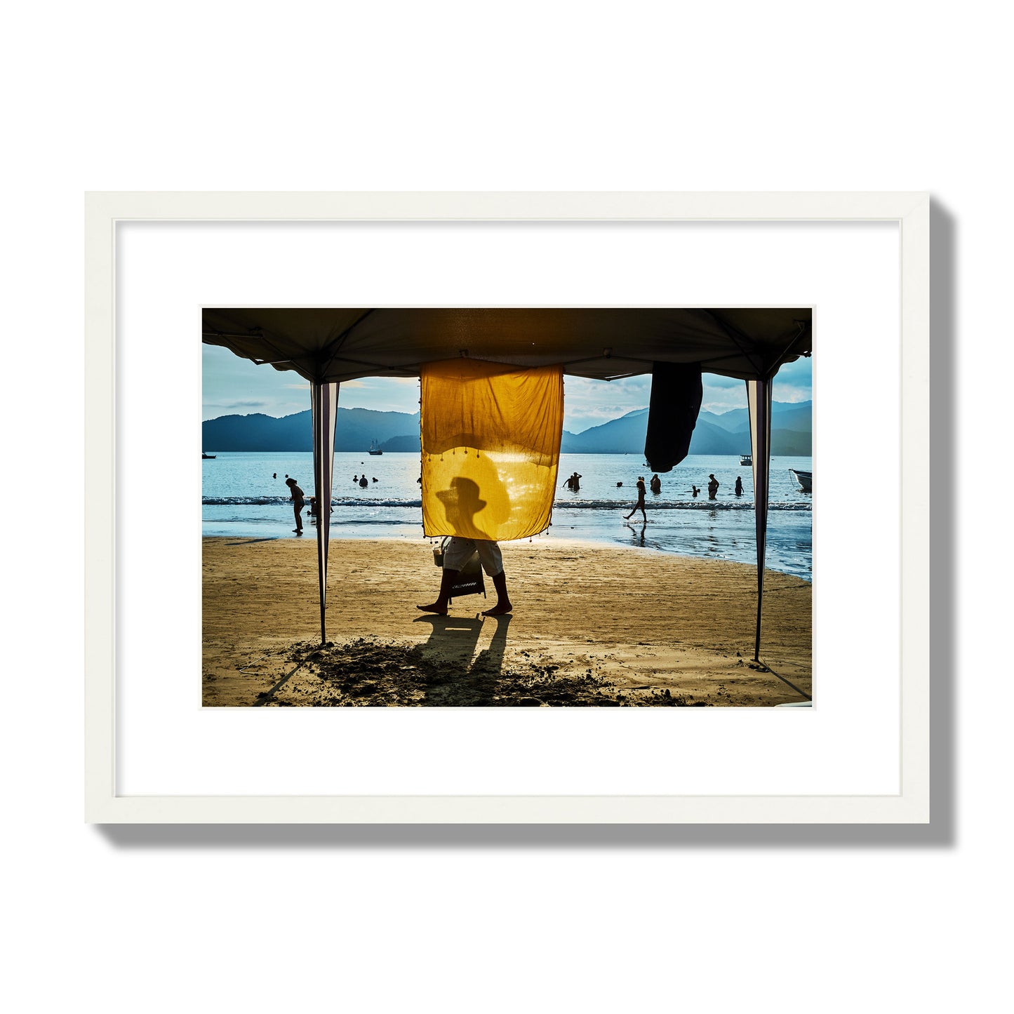 Fine art street photograph of a crowded beach scene with a vibrant yellow flap hanging from a tent, sunlight filtering through and silhouetting a man’s face. Archival pigment print, medium size, white frame.
