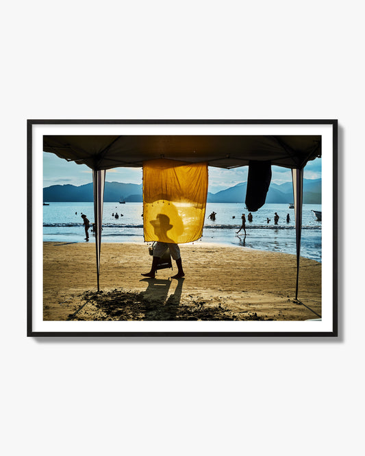 Fine art street photograph of a crowded beach scene with a vibrant yellow flap hanging from a tent, sunlight filtering through and silhouetting a man’s face. Archival pigment print, black frame.