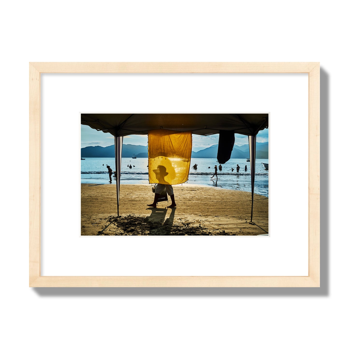 Fine art street photograph of a crowded beach scene with a vibrant yellow flap hanging from a tent, sunlight filtering through and silhouetting a man’s face. Archival pigment print, small size, natural wood frame.