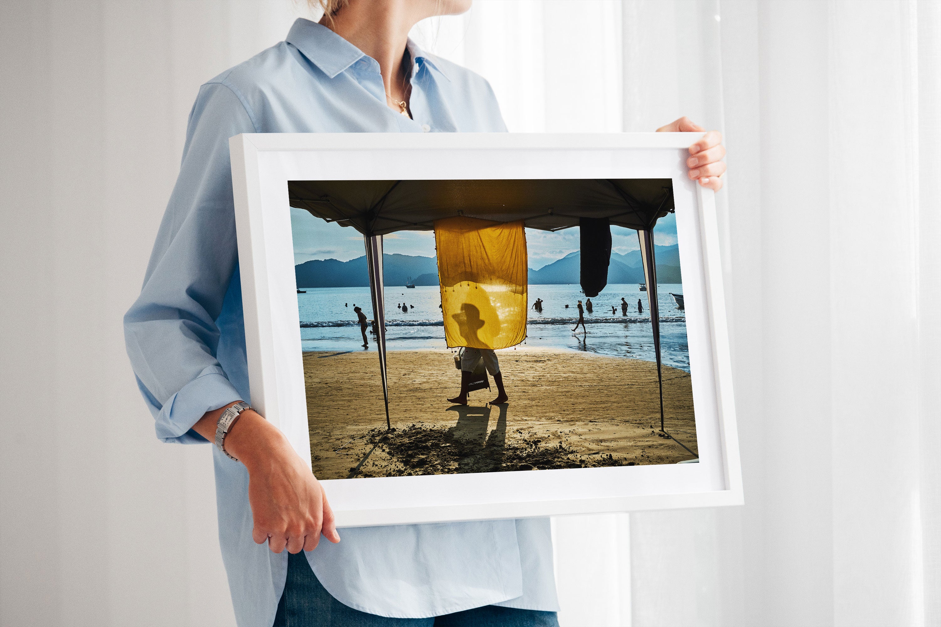 A woman holding a white-framed archival pigment print depicting a man walking in a beach scene.