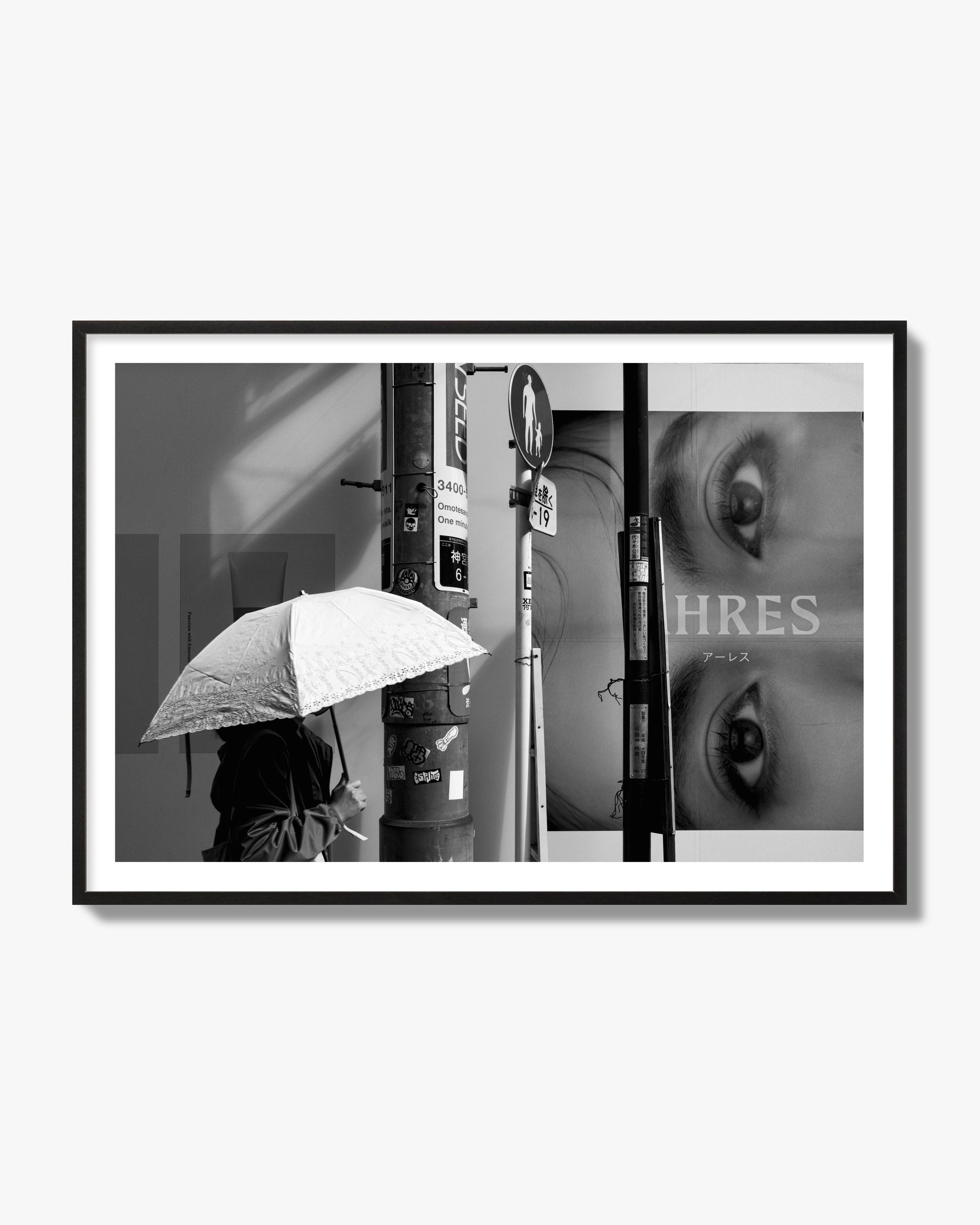 Fine art street photograph of a woman walking with an umbrella on a Tokyo street, passing beneath a billboard with two large eyes. Archival pigment print, black frame.