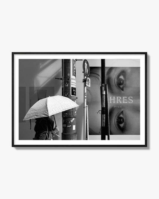Fine art street photograph of a woman walking with an umbrella on a Tokyo street, passing beneath a billboard with two large eyes. Archival pigment print, black frame.