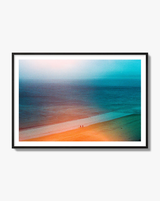 Fine art street photograph of a serene beach with two figures walking in the distance, captured in a dreamlike composition with soft orange and blue hues. Archival pigment print, black frame.