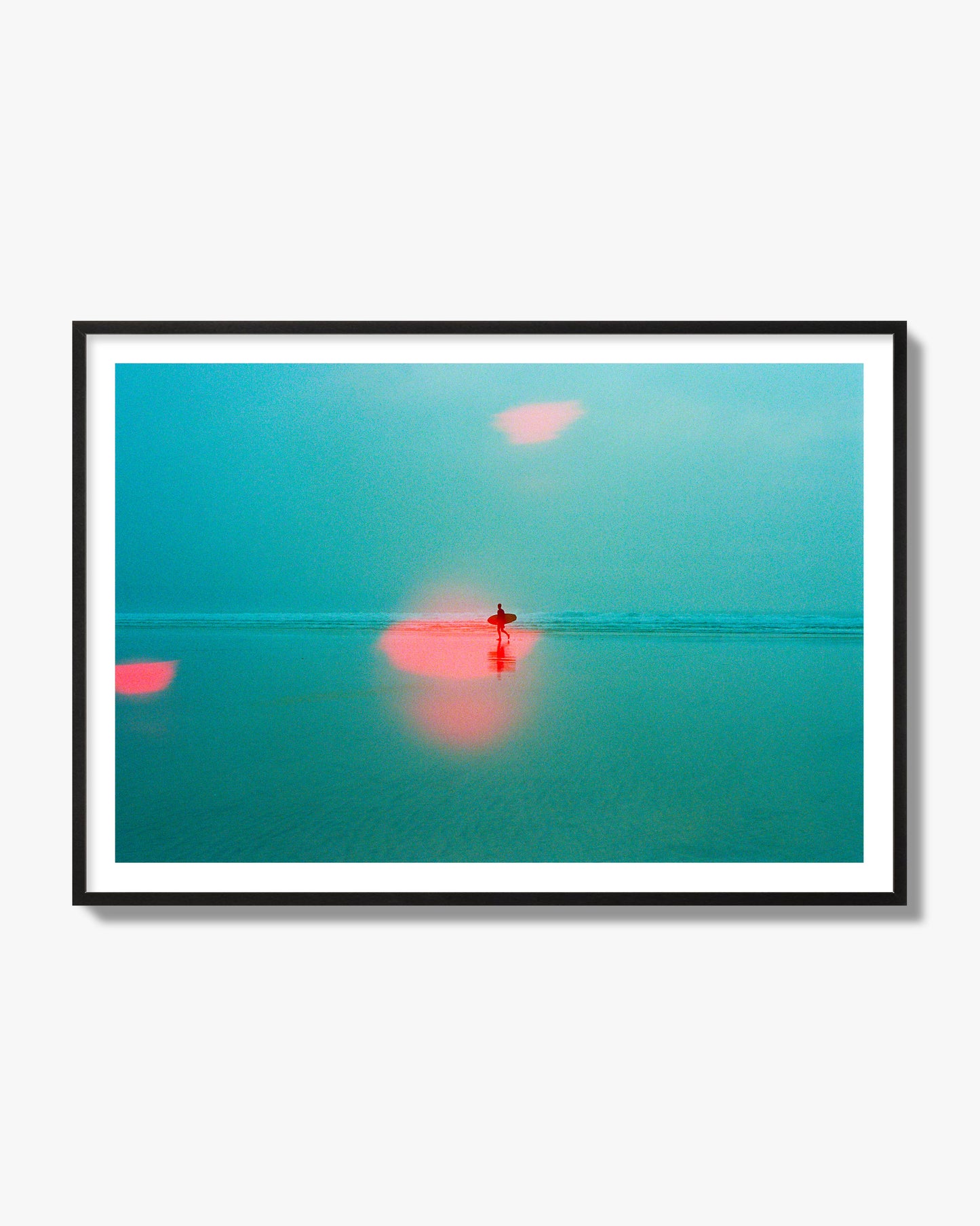 Fine art street photograph of a tranquil beach with a surfer running toward the waves, featuring a dreamlike composition with soft teal tones and a striking red lens flare. Archival pigment print, black frame.