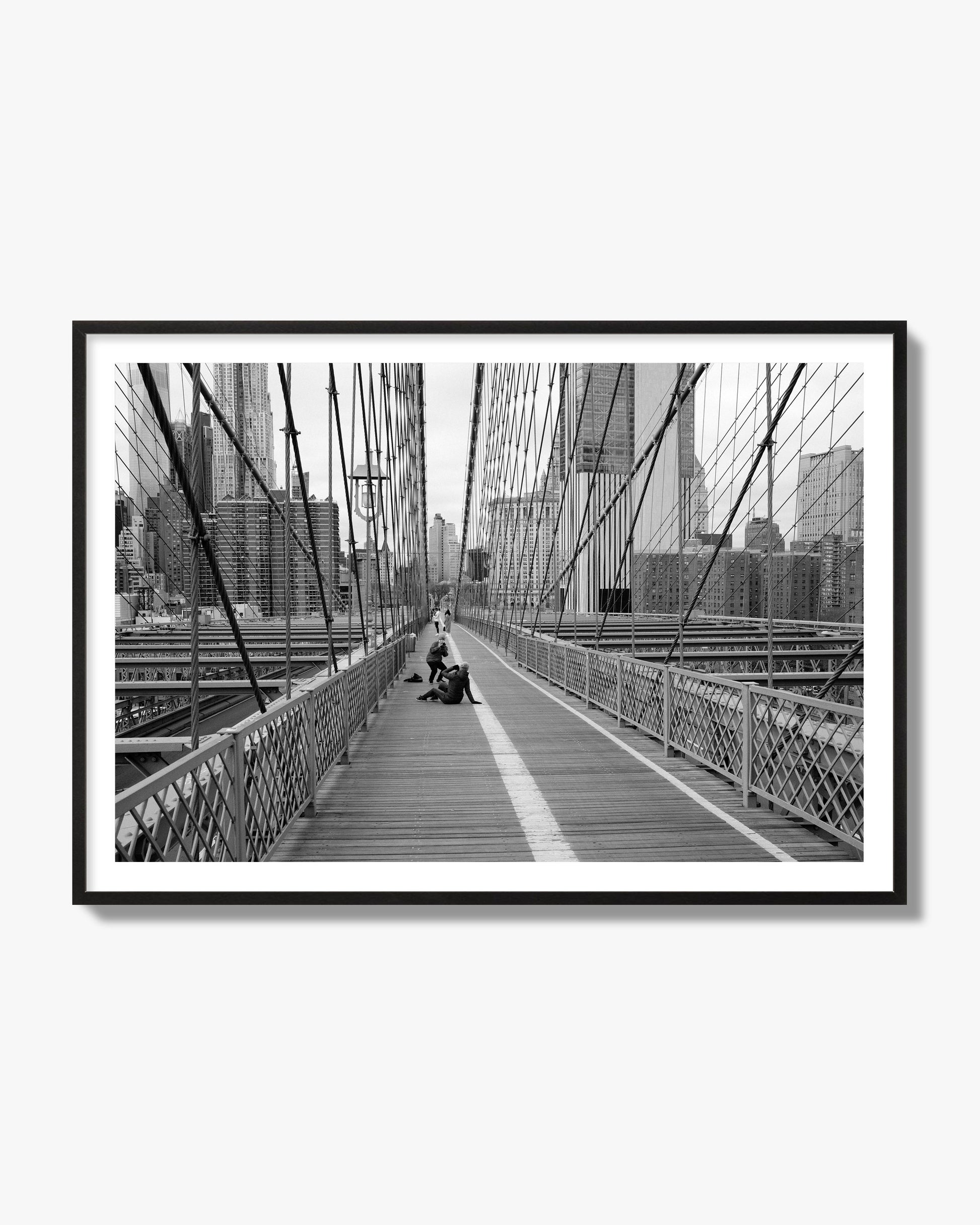 Fine art black and white photograph of a person posing on the Brooklyn Bridge with the cityscape in the background, archival pigment print, black frame
