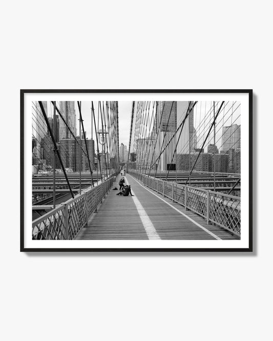 Fine art black and white photograph of a person posing on the Brooklyn Bridge with the cityscape in the background, archival pigment print, black frame