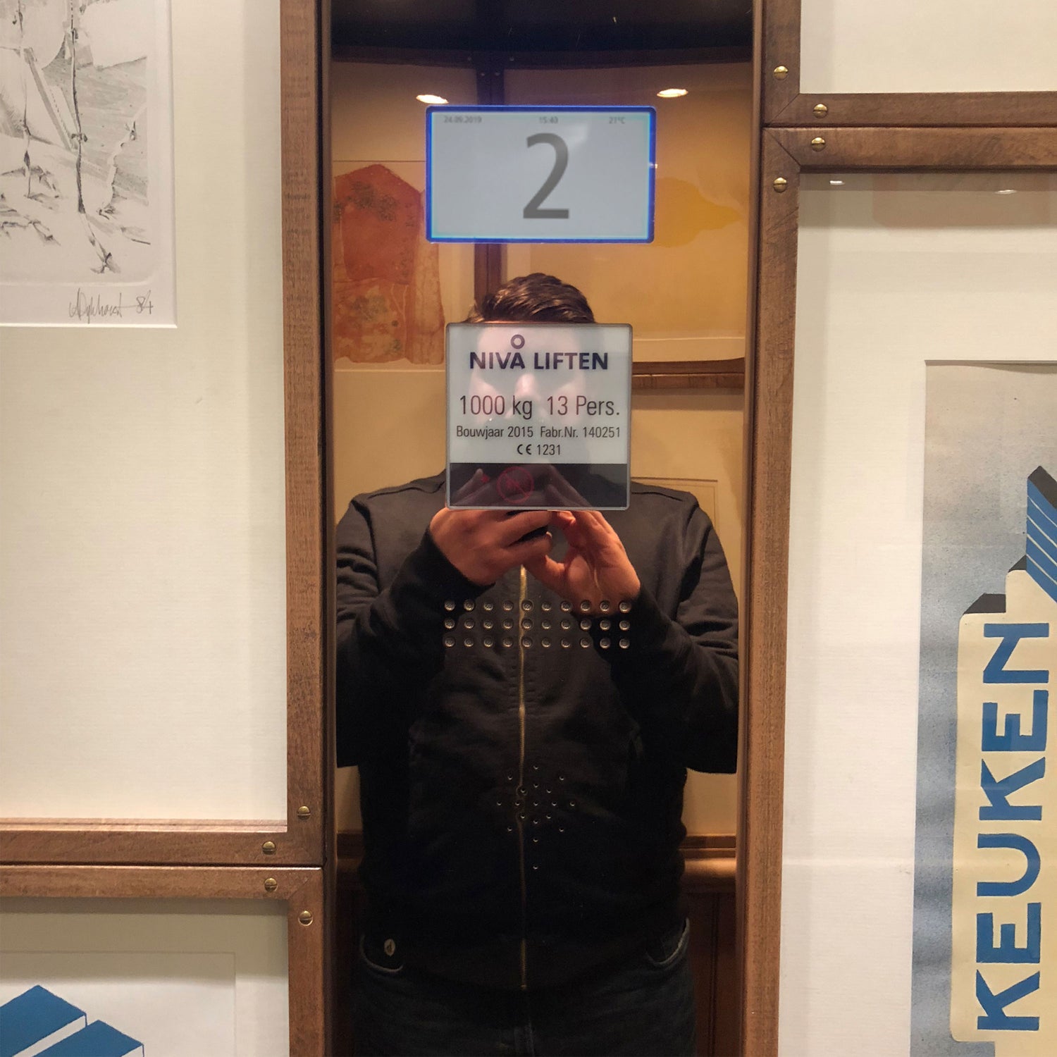 A reflection of a man in an elevator mirror, his face obscured by the writing on the elevator’s digital display.