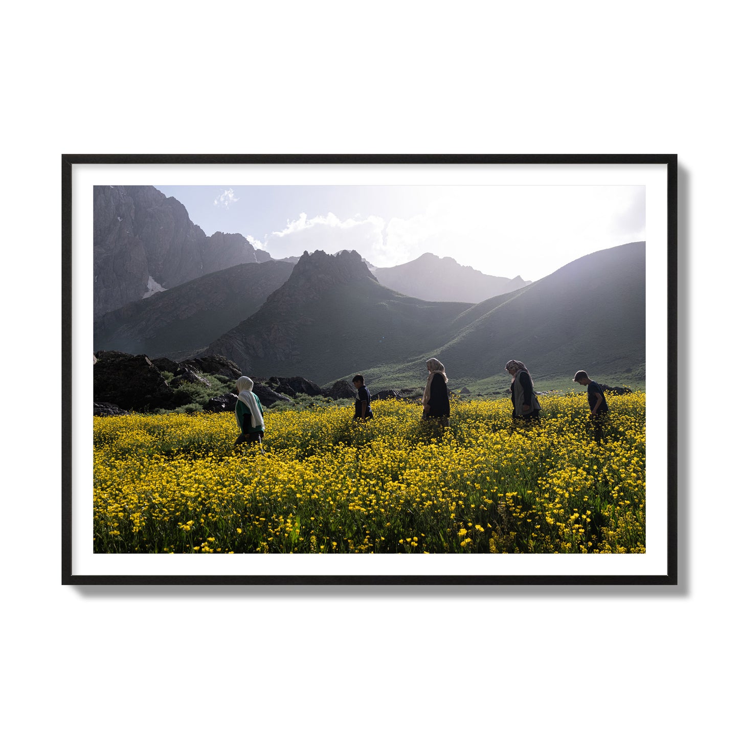 Fine art photograph of people walking through a field of yellow flowers with mountains in the background, archival pigment print, collector size, black frame.