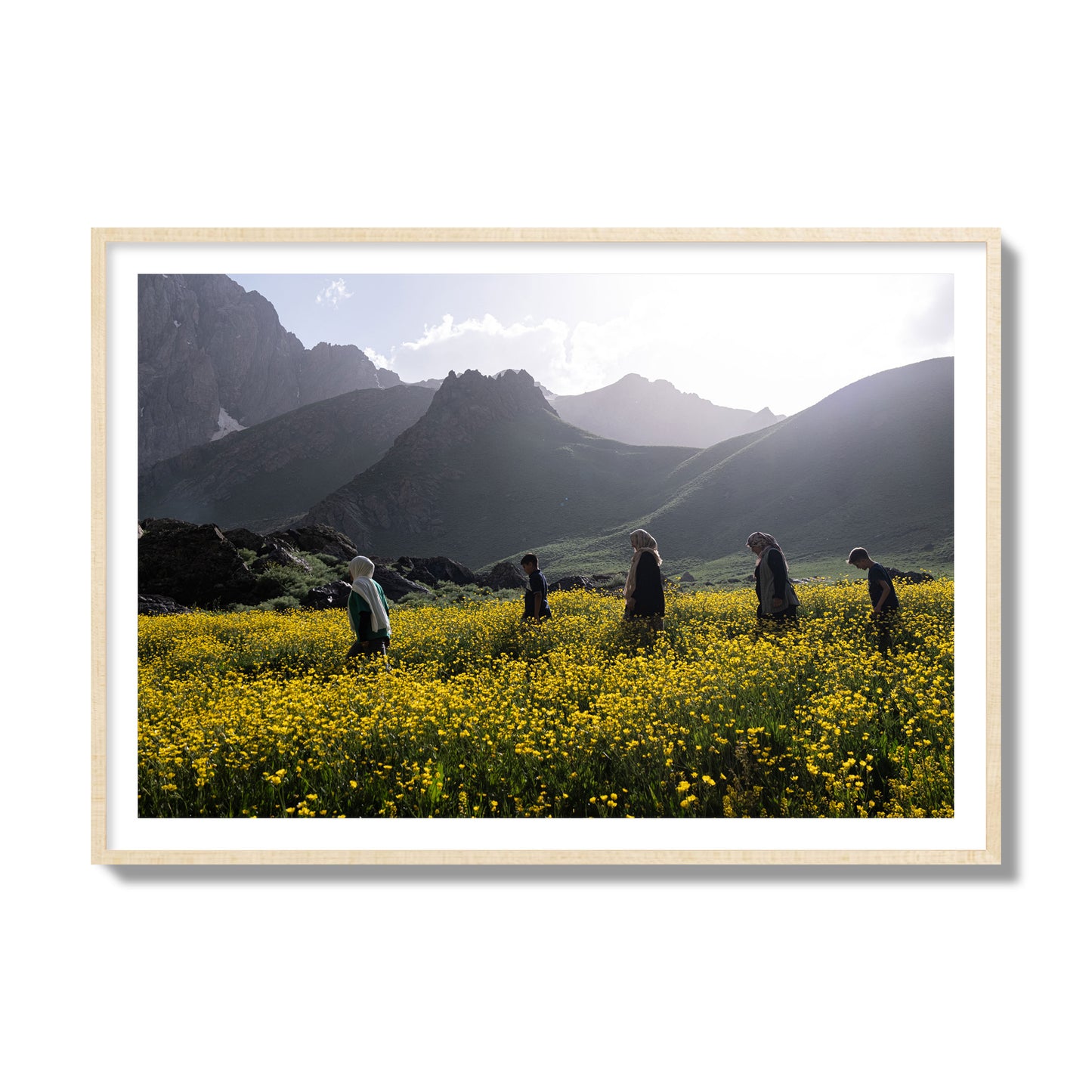 Fine art photograph of people walking through a field of yellow flowers with mountains in the background, archival pigment print, collector size, natural wood frame.