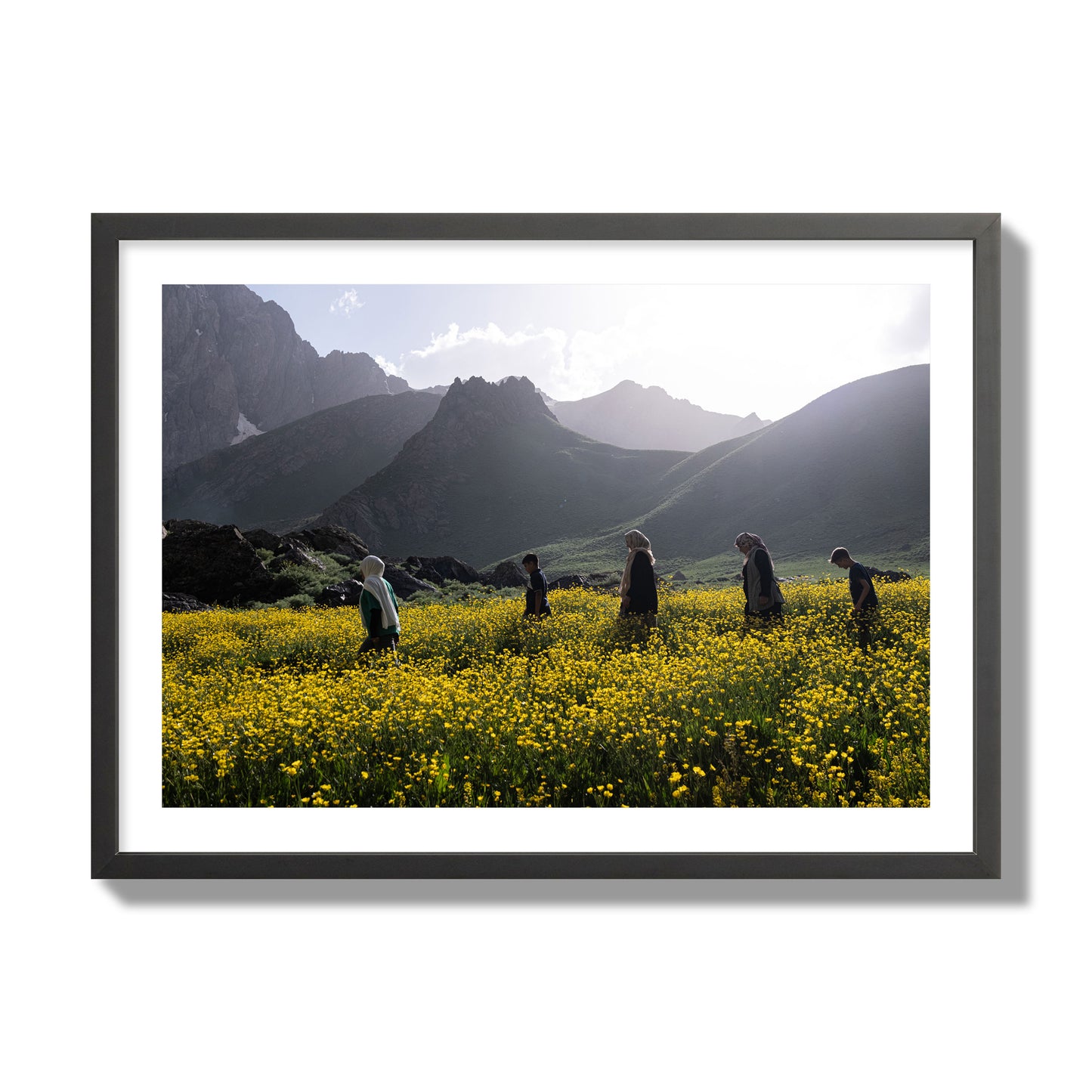 Fine art photograph of people walking through a field of yellow flowers with mountains in the background, archival pigment print, giant size, black frame.