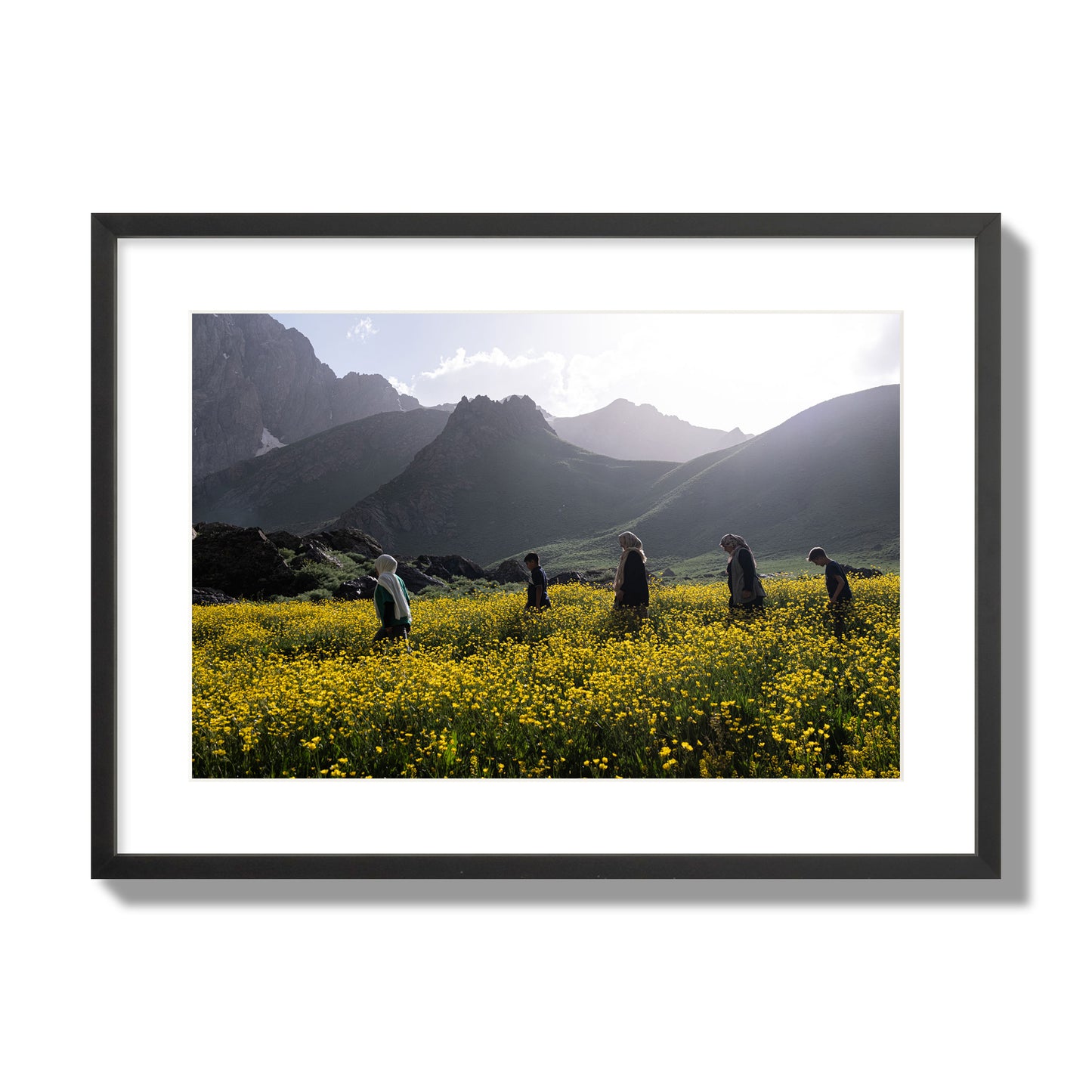 Fine art photograph of people walking through a field of yellow flowers with mountains in the background, archival pigment print, large size, black frame.