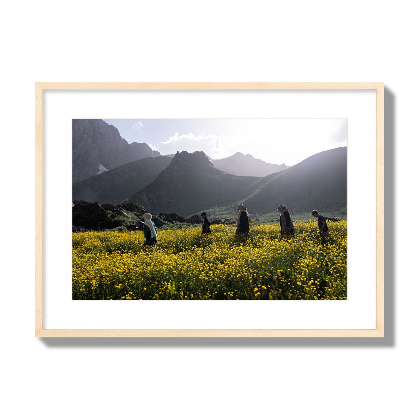 Fine art photograph of people walking through a field of yellow flowers with mountains in the background, archival pigment print, large size, natural wood frame.