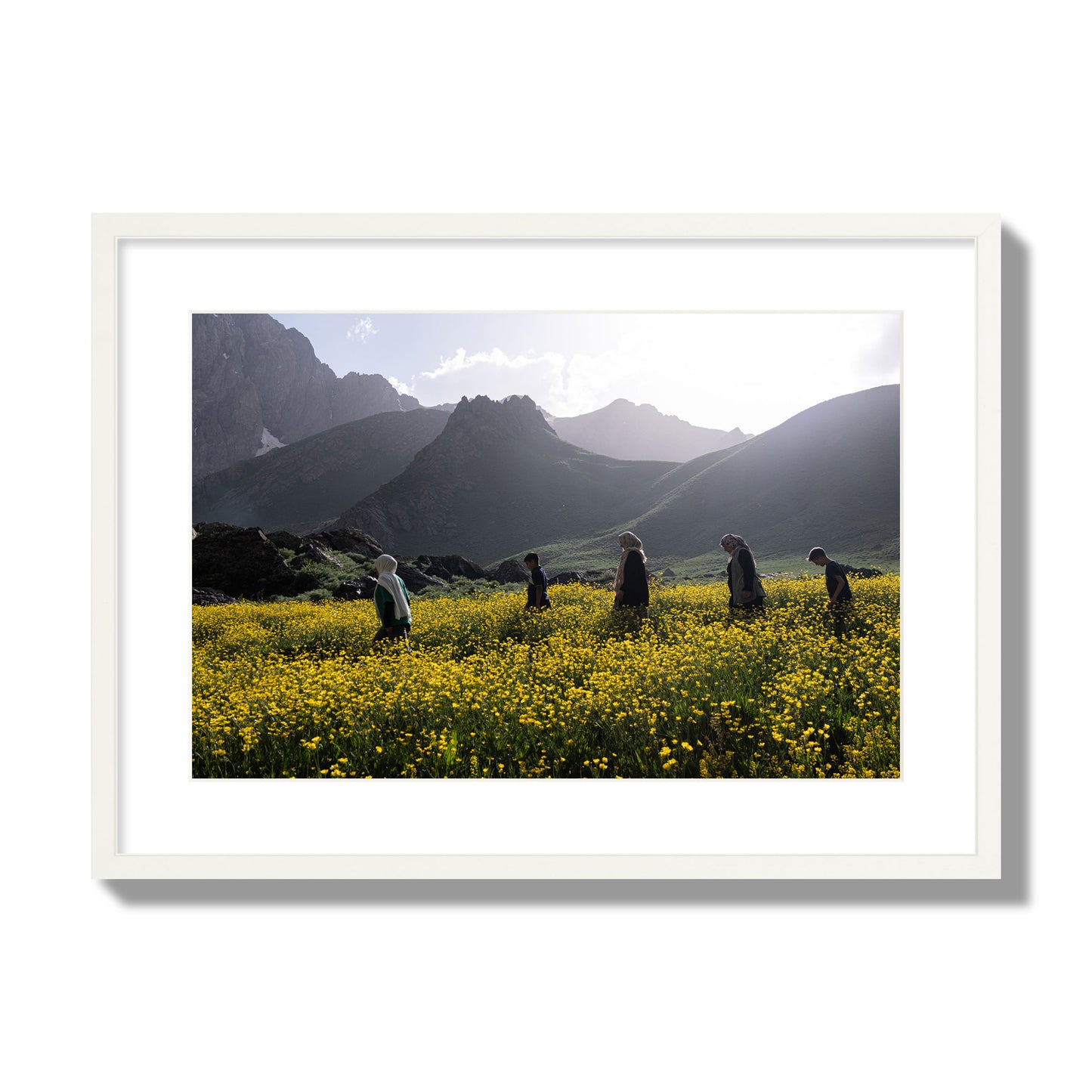 Fine art photograph of people walking through a field of yellow flowers with mountains in the background, archival pigment print, large size, white frame.