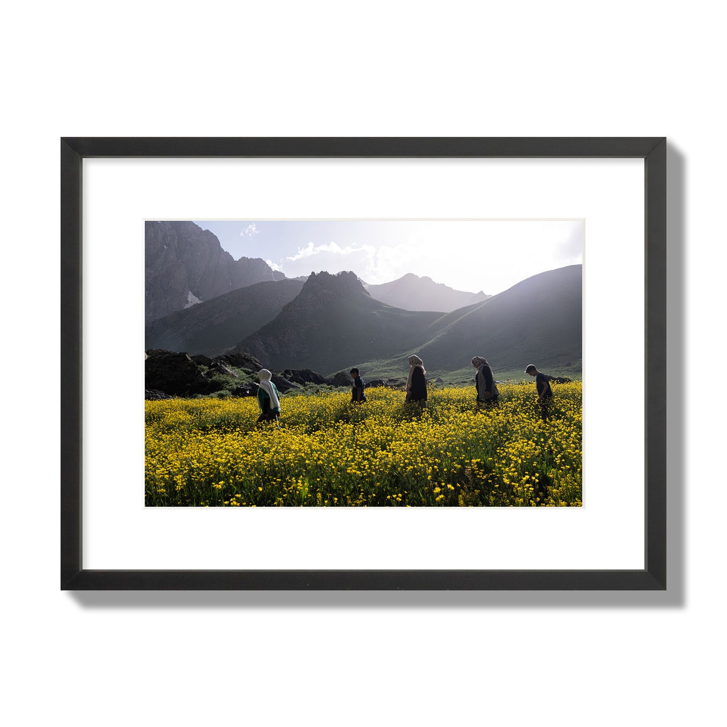 Fine art photograph of people walking through a field of yellow flowers with mountains in the background, archival pigment print, medium size, black frame.
