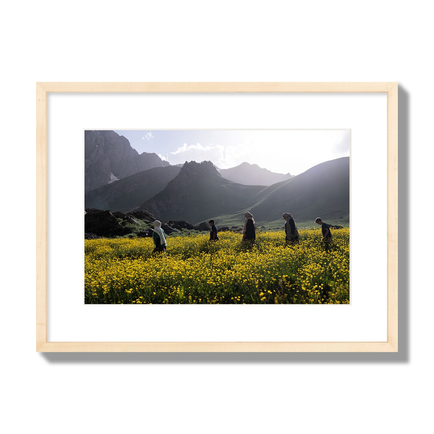 Fine art photograph of people walking through a field of yellow flowers with mountains in the background, archival pigment print, medium size, natural wood frame.
