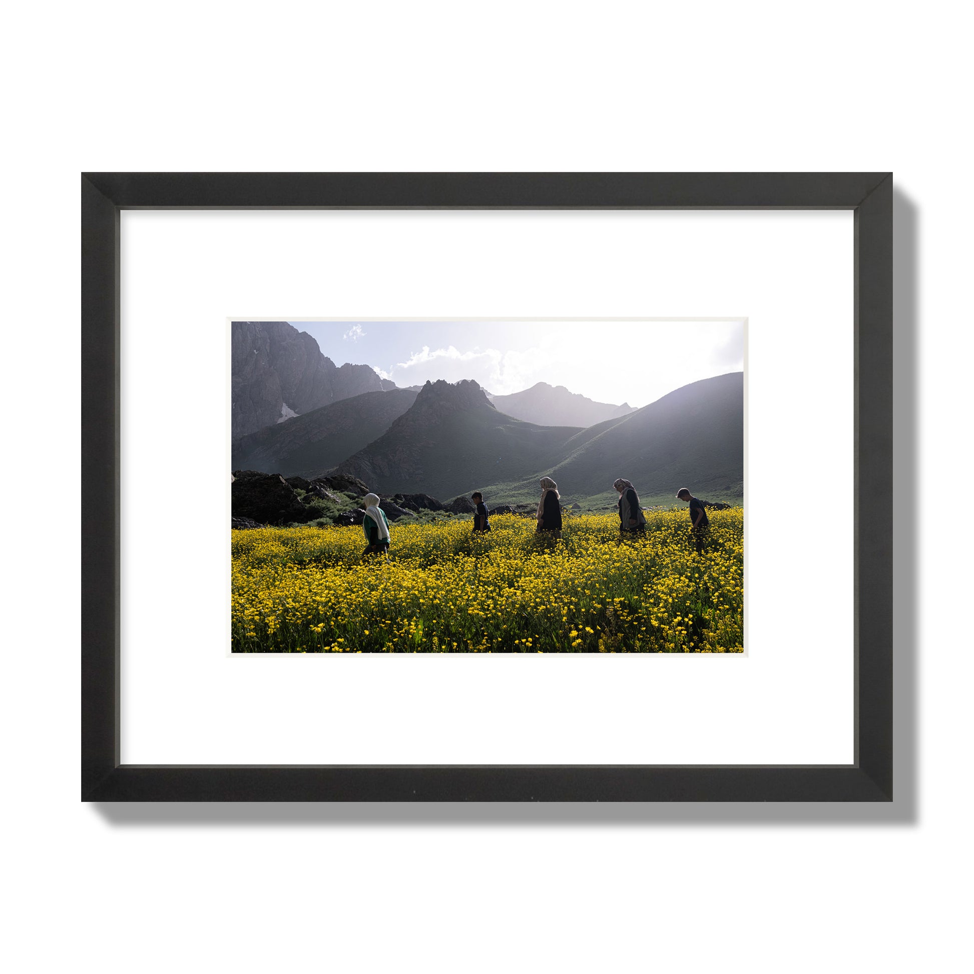 Fine art photograph of people walking through a field of yellow flowers with mountains in the background, archival pigment print, small size, black frame.