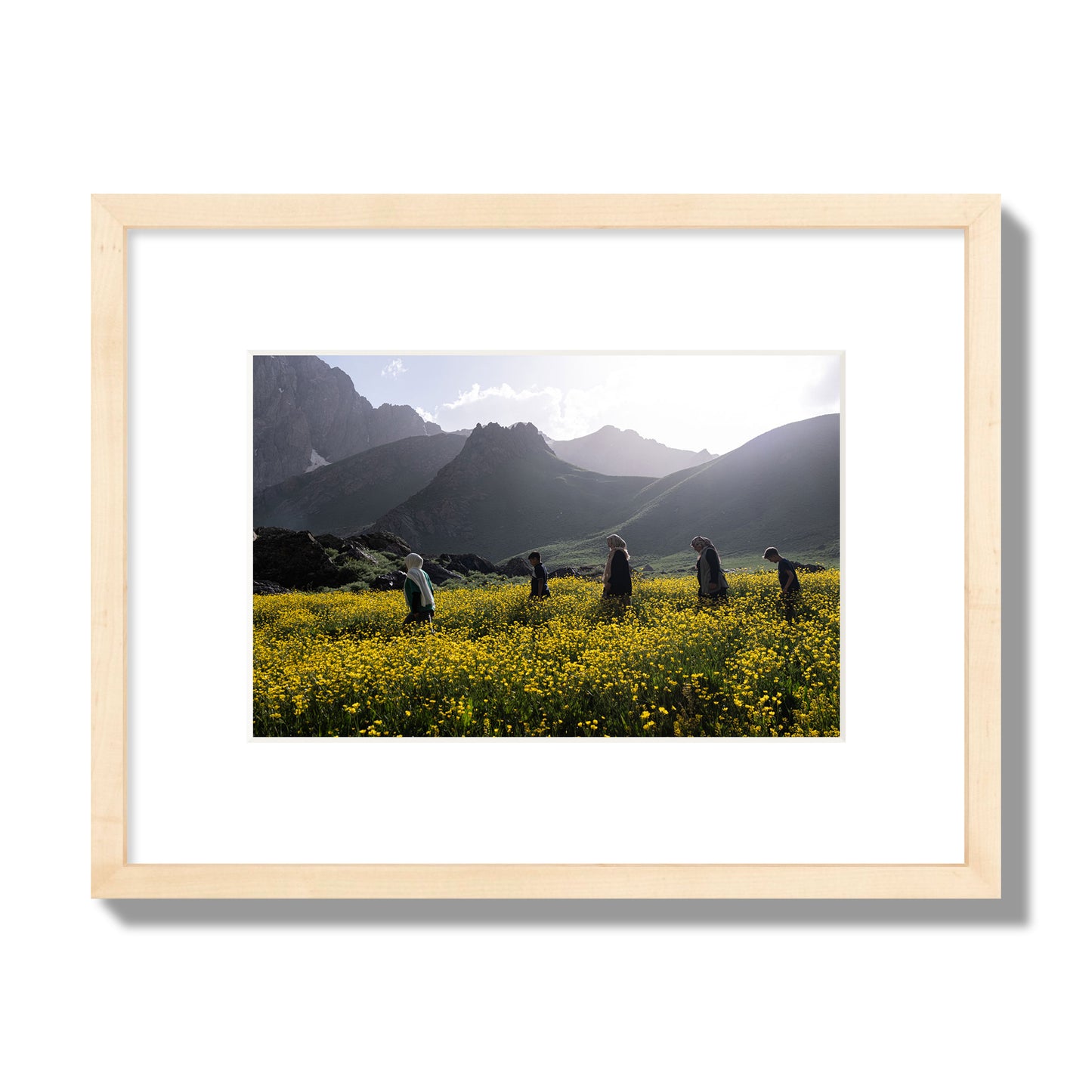 Fine art photograph of people walking through a field of yellow flowers with mountains in the background, archival pigment print, small size, natural wood frame.