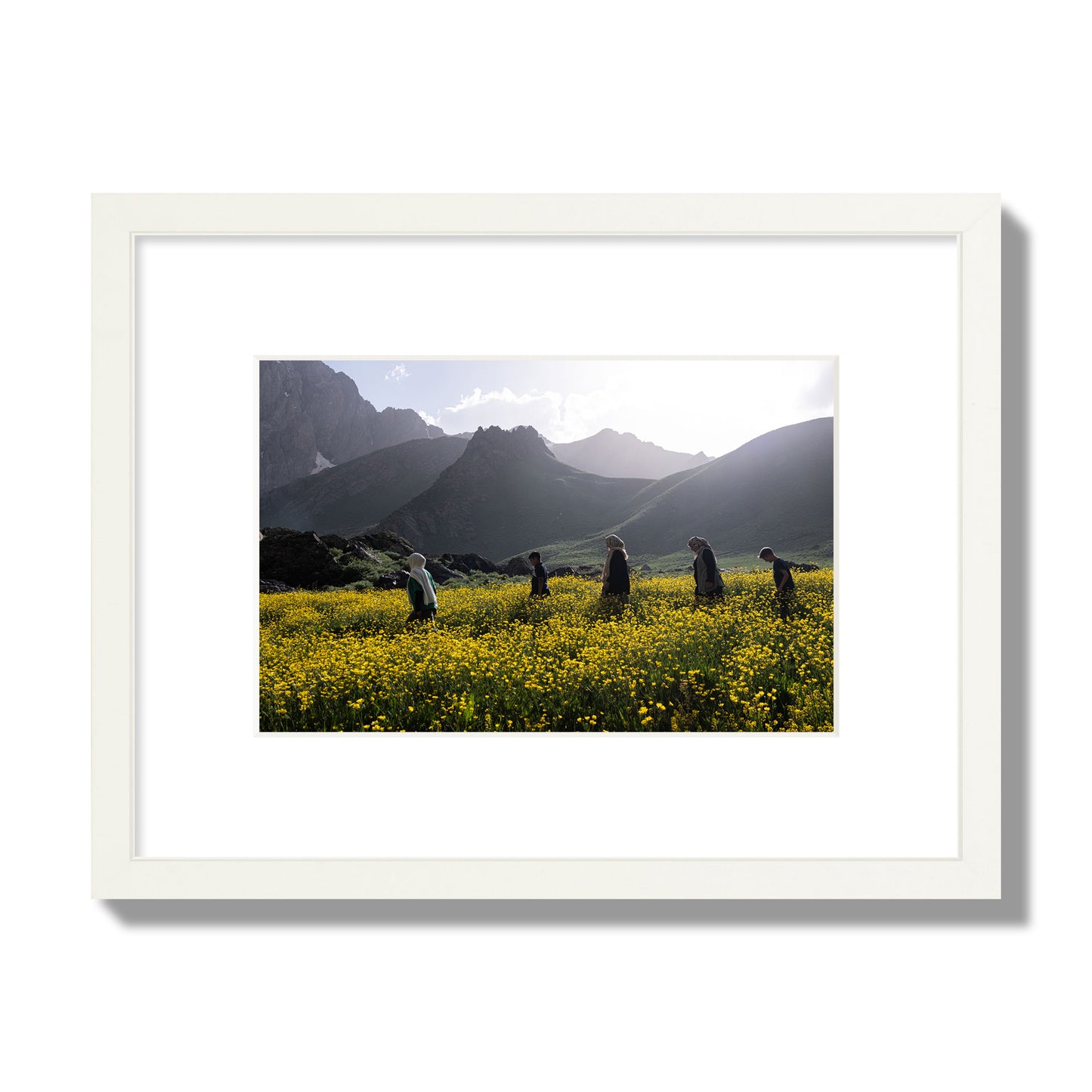 Fine art photograph of people walking through a field of yellow flowers with mountains in the background, archival pigment print, small size, white frame.