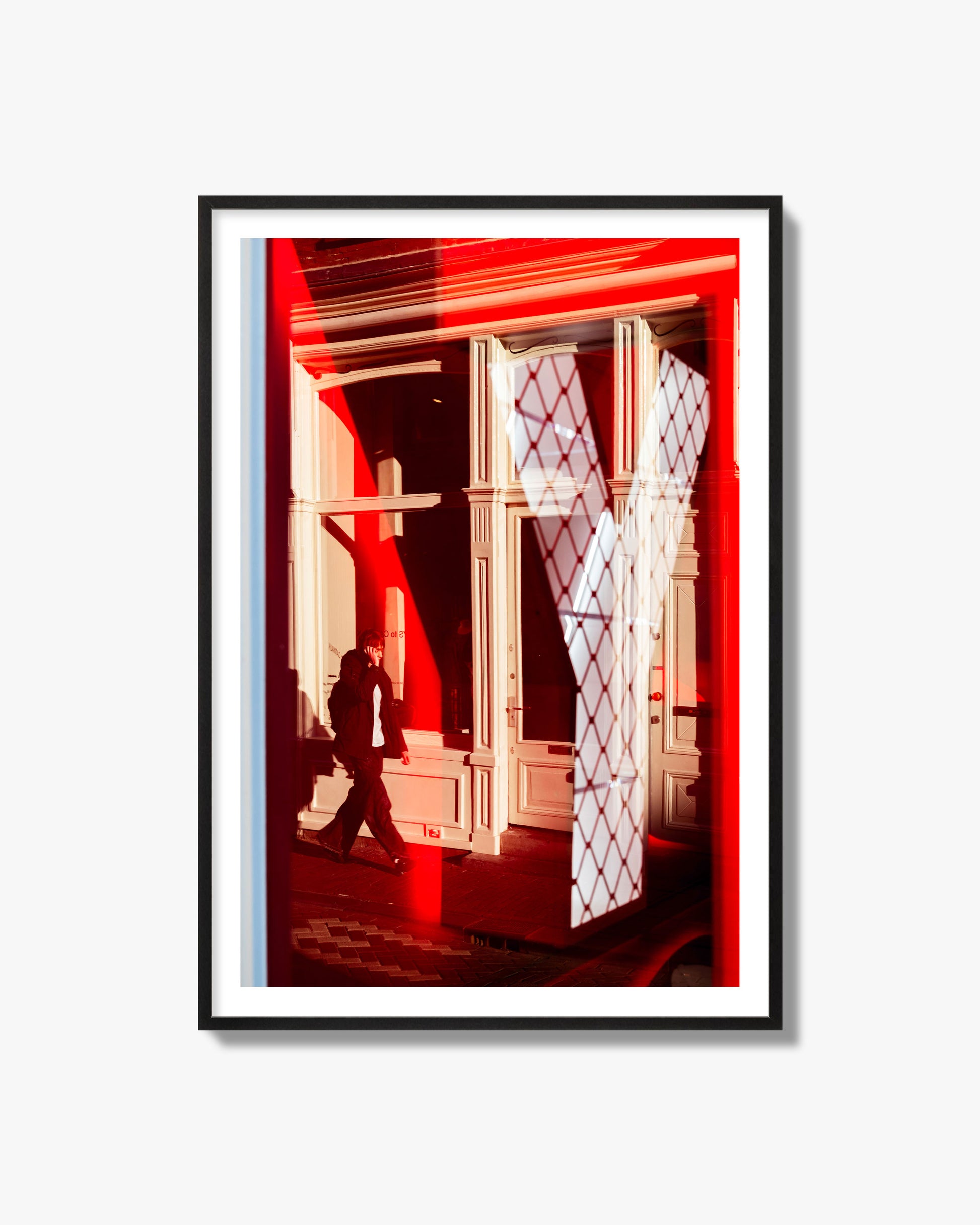 Fine art street photograph of an Amsterdam street scene showing bright red and white reflections of a large letter Y, a man walking, and multiple store fronts. Archival pigment print, black frame.