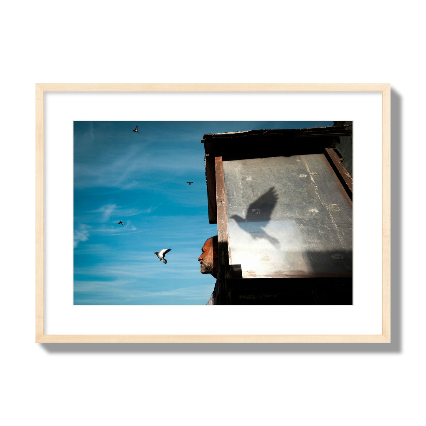 Fine art street photograph of a man watching a bird fly away in the background, with the bird’s shadow cast across a birdcage in the foreground. Archival pigment print, large size, natural wood frame.