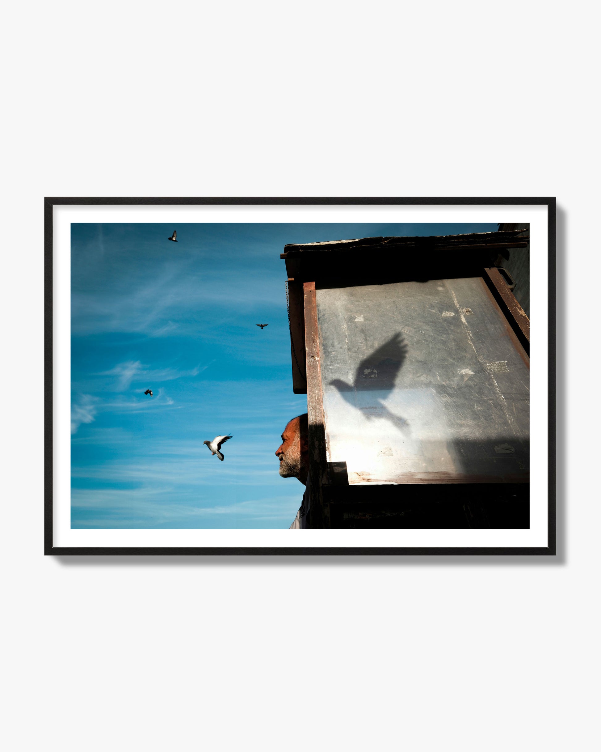 Fine art street photograph of a man watching a bird fly away in the background, with the bird’s shadow cast across a birdcage in the foreground. Archival pigment print, black frame.