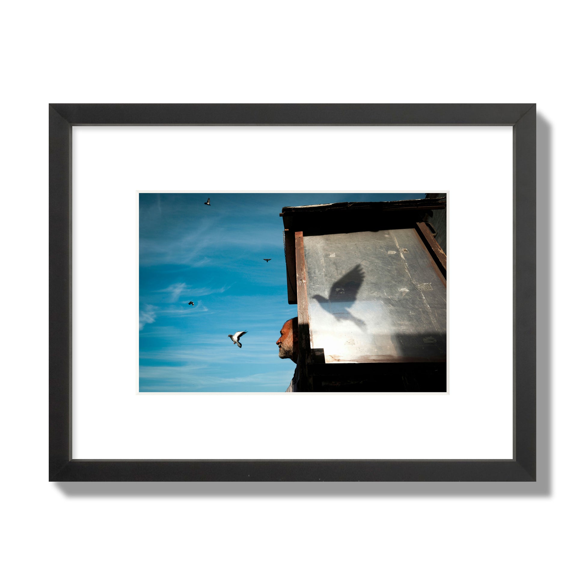 Fine art street photograph of a man watching a bird fly away in the background, with the bird’s shadow cast across a birdcage in the foreground. Archival pigment print, small size, black frame.