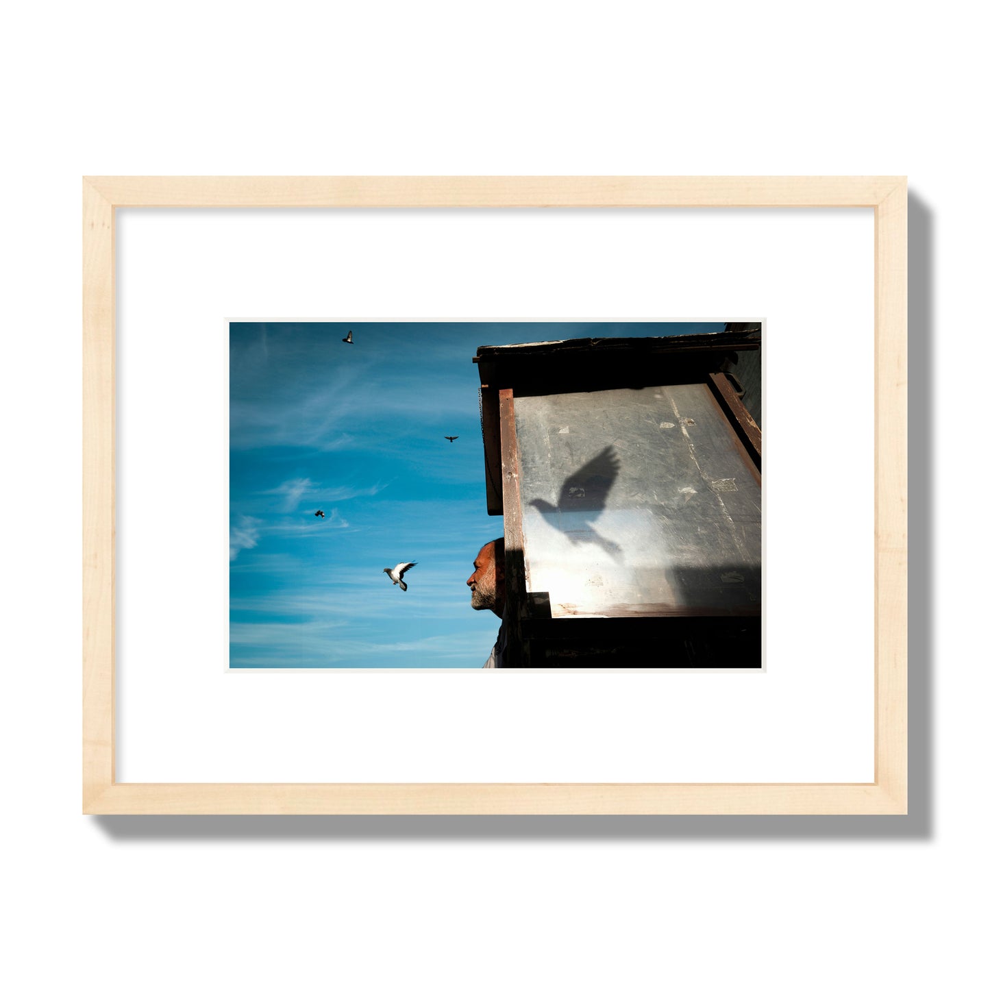 Fine art street photograph of a man watching a bird fly away in the background, with the bird’s shadow cast across a birdcage in the foreground. Archival pigment print, small size, natural wood frame.