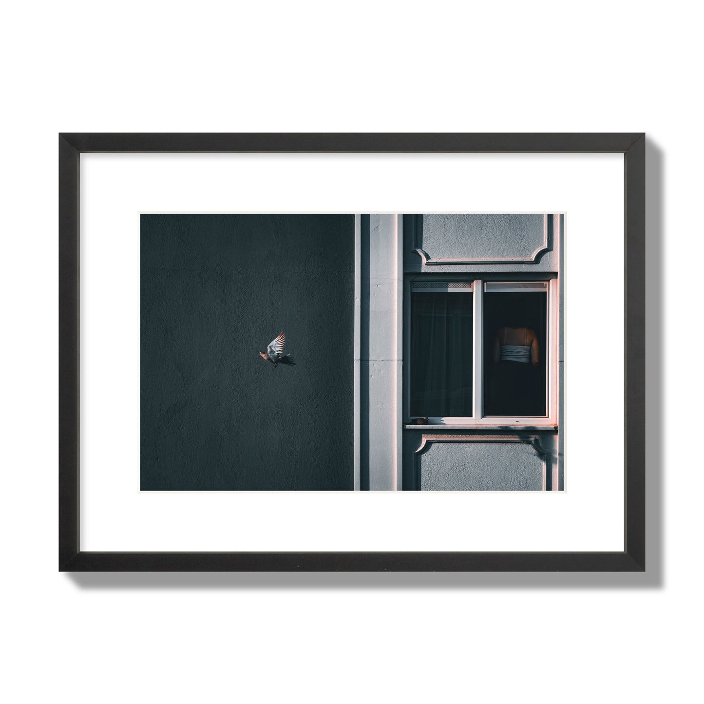Fine art street photograph of a woman highlighted in a window on the right side of the frame, while a bird flies out of frame on the left. Archival pigment print, medium size, black frame.