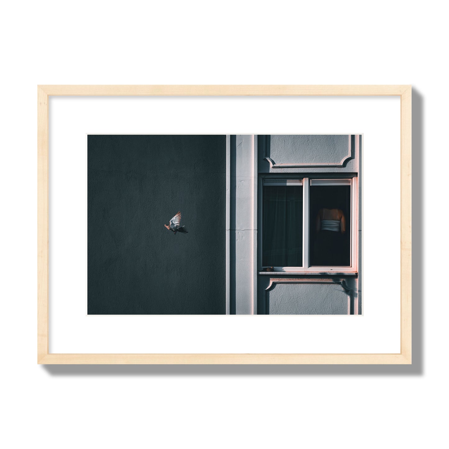 Fine art street photograph of a woman highlighted in a window on the right side of the frame, while a bird flies out of frame on the left. Archival pigment print, medium size, natural wood frame.
