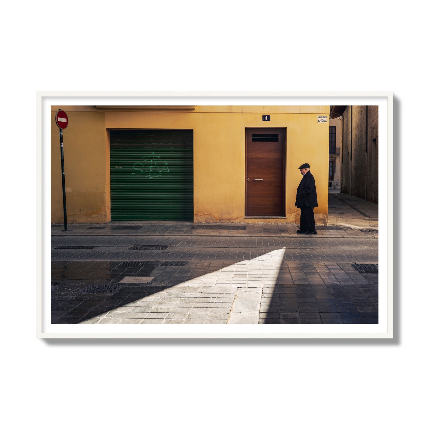 Fine art street photograph of an elderly man walking down a Valencia, Spain street with yellow and green walls, illuminated by a shaft of light. Archival pigment print, gallery size, white frame.