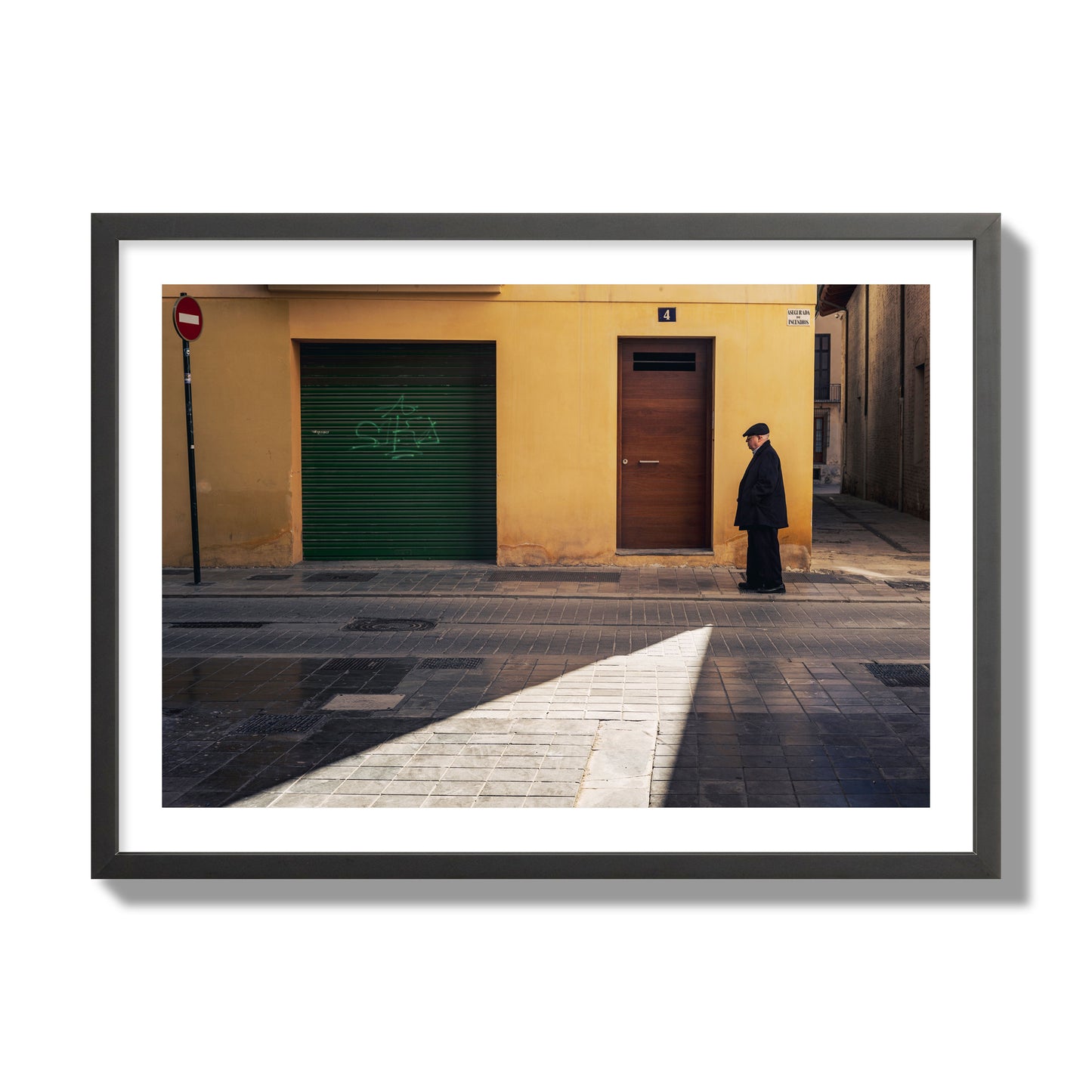 Fine art street photograph of an elderly man walking down a Valencia, Spain street with yellow and green walls, illuminated by a shaft of light. Archival pigment print, giant size, black frame.