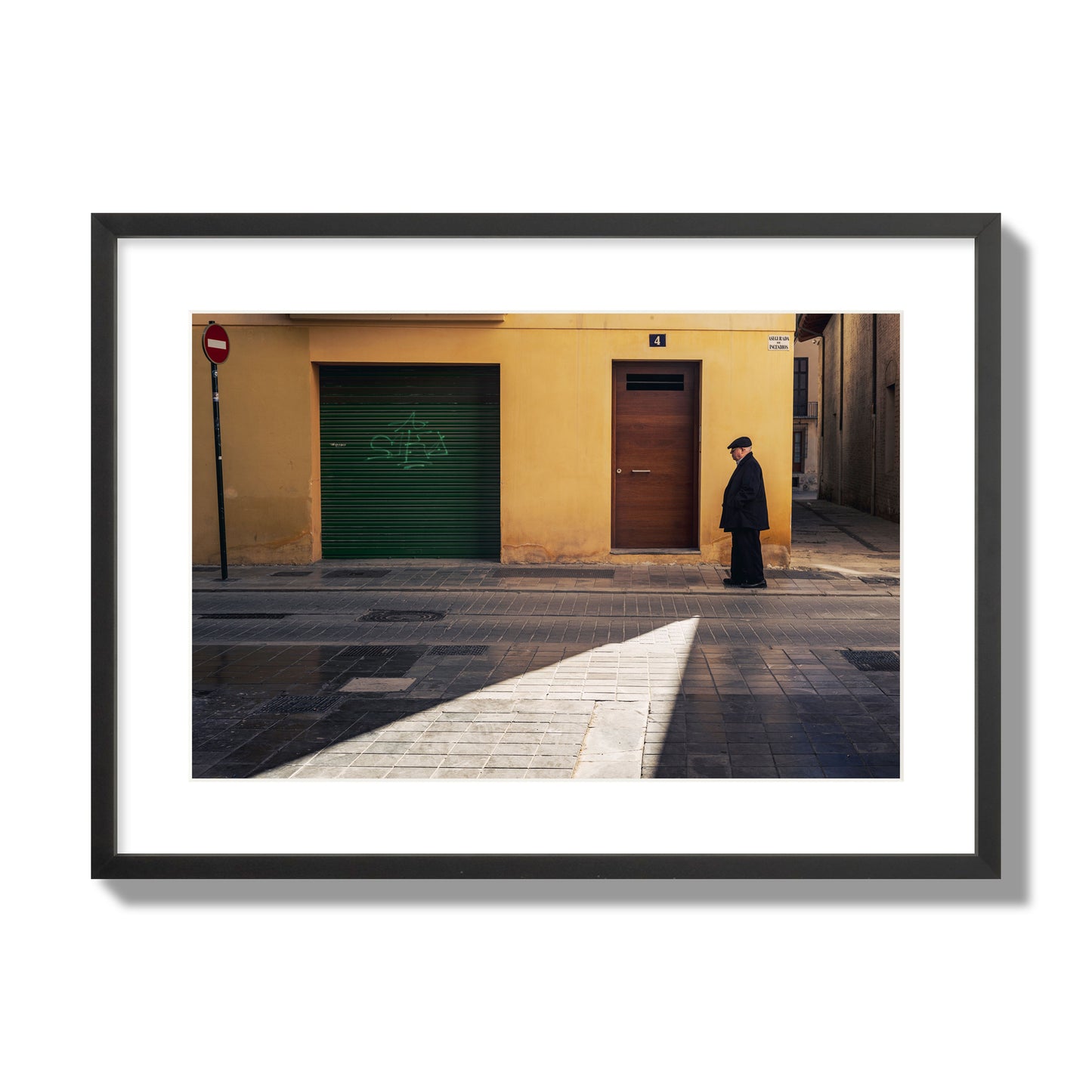 Fine art street photograph of an elderly man walking down a Valencia, Spain street with yellow and green walls, illuminated by a shaft of light. Archival pigment print, large size, black frame.