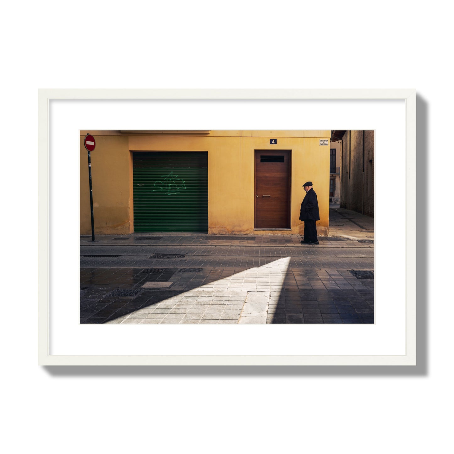 Fine art street photograph of an elderly man walking down a Valencia, Spain street with yellow and green walls, illuminated by a shaft of light. Archival pigment print, large size, white frame.