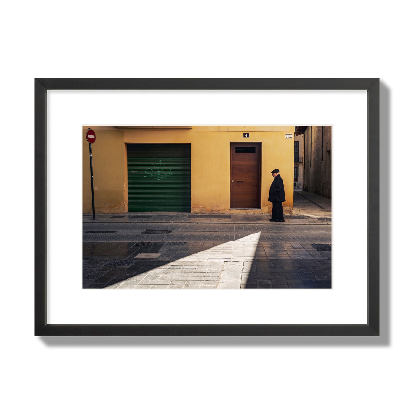 Fine art street photograph of an elderly man walking down a Valencia, Spain street with yellow and green walls, illuminated by a shaft of light. Archival pigment print, medium size, black frame.