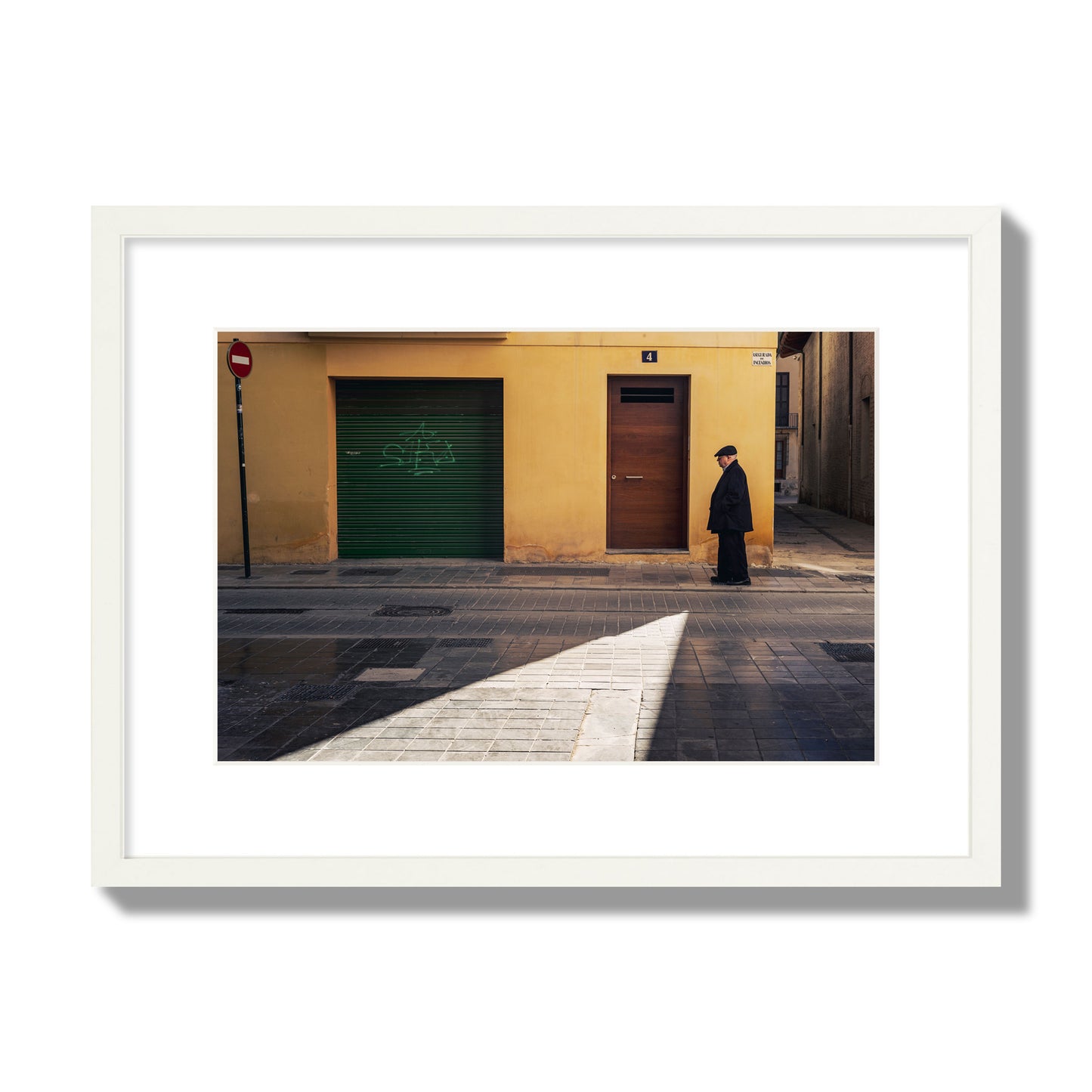 Fine art street photograph of an elderly man walking down a Valencia, Spain street with yellow and green walls, illuminated by a shaft of light. Archival pigment print, medium size, white frame.