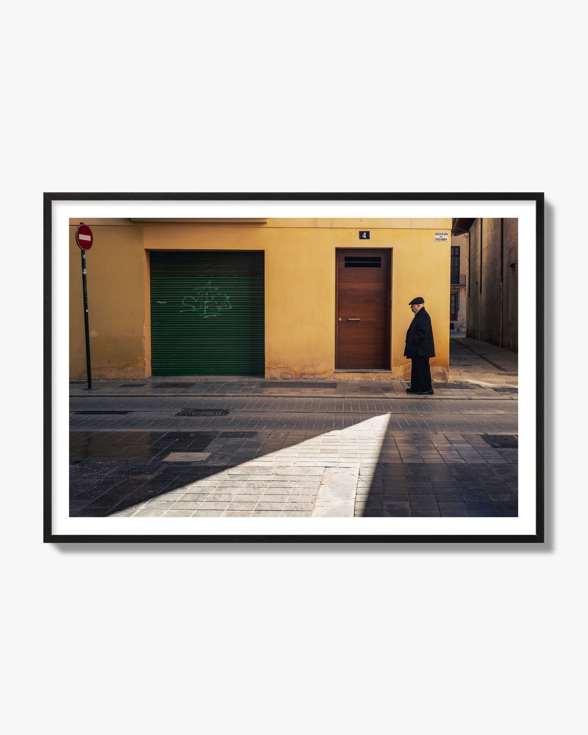 Fine art street photograph of an elderly man walking down a Valencia, Spain street with yellow and green walls, illuminated by a shaft of light. Archival pigment print, black frame.