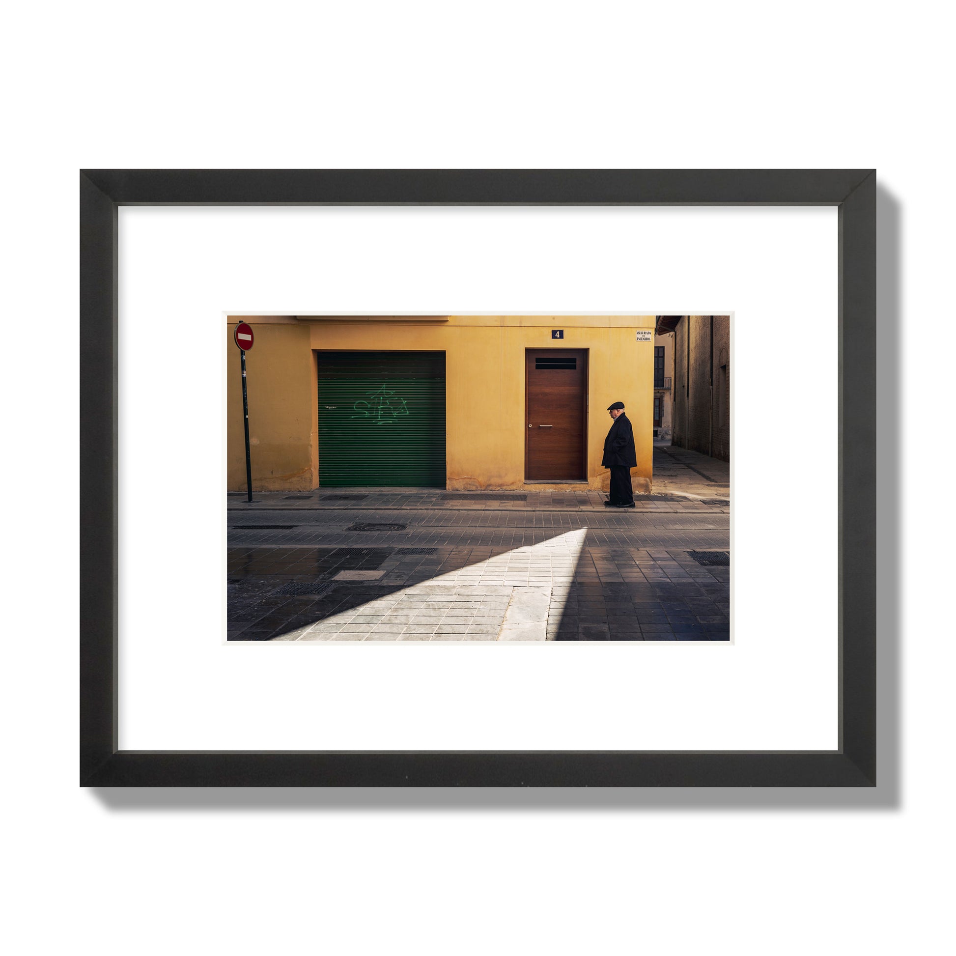 Fine art street photograph of an elderly man walking down a Valencia, Spain street with yellow and green walls, illuminated by a shaft of light. Archival pigment print, small size, black frame.