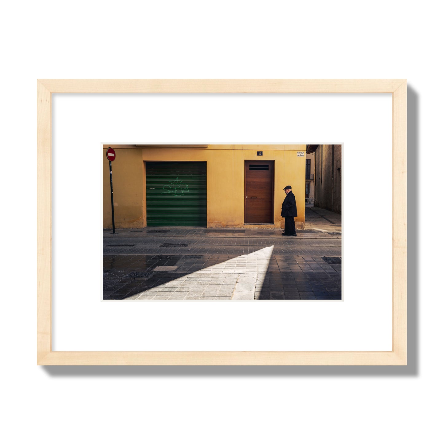 Fine art street photograph of an elderly man walking down a Valencia, Spain street with yellow and green walls, illuminated by a shaft of light. Archival pigment print, small size, natural wood frame.