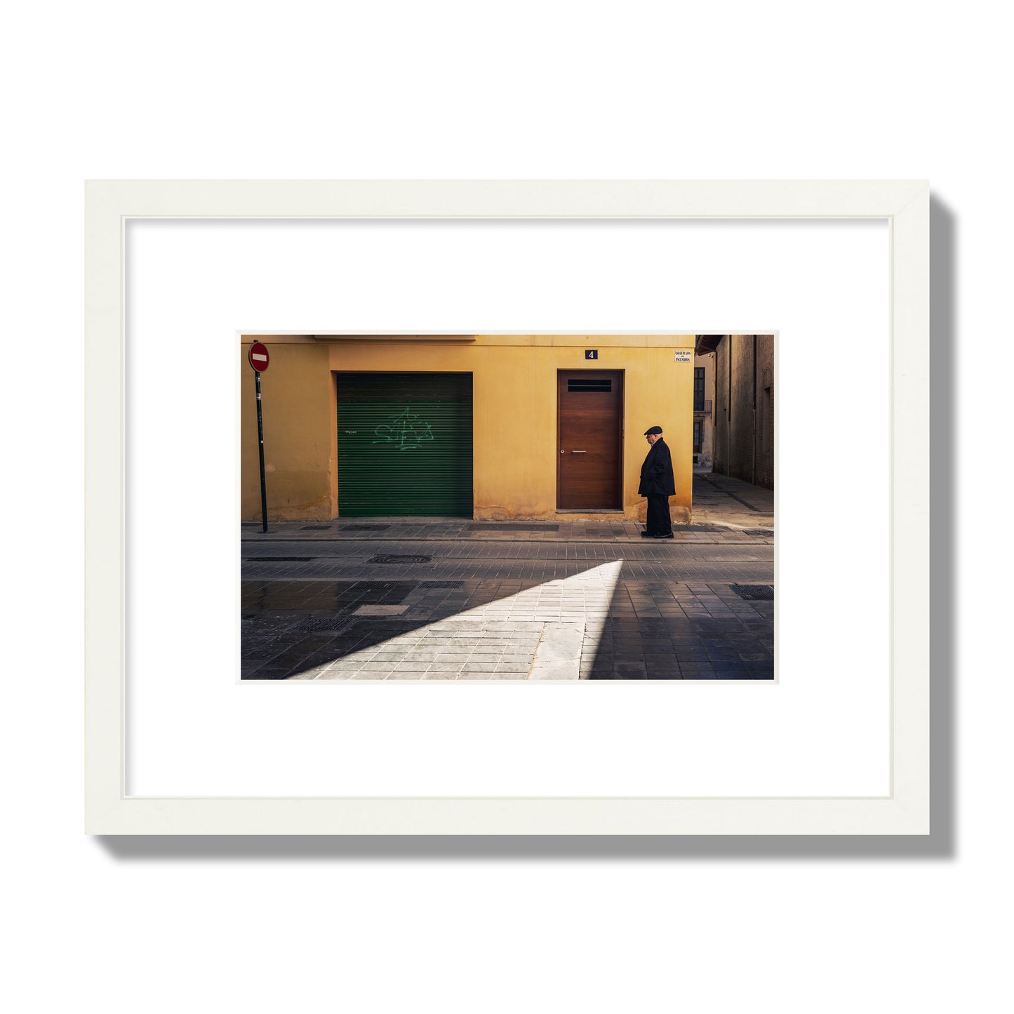 Fine art street photograph of an elderly man walking down a Valencia, Spain street with yellow and green walls, illuminated by a shaft of light. Archival pigment print, small size, white frame.
